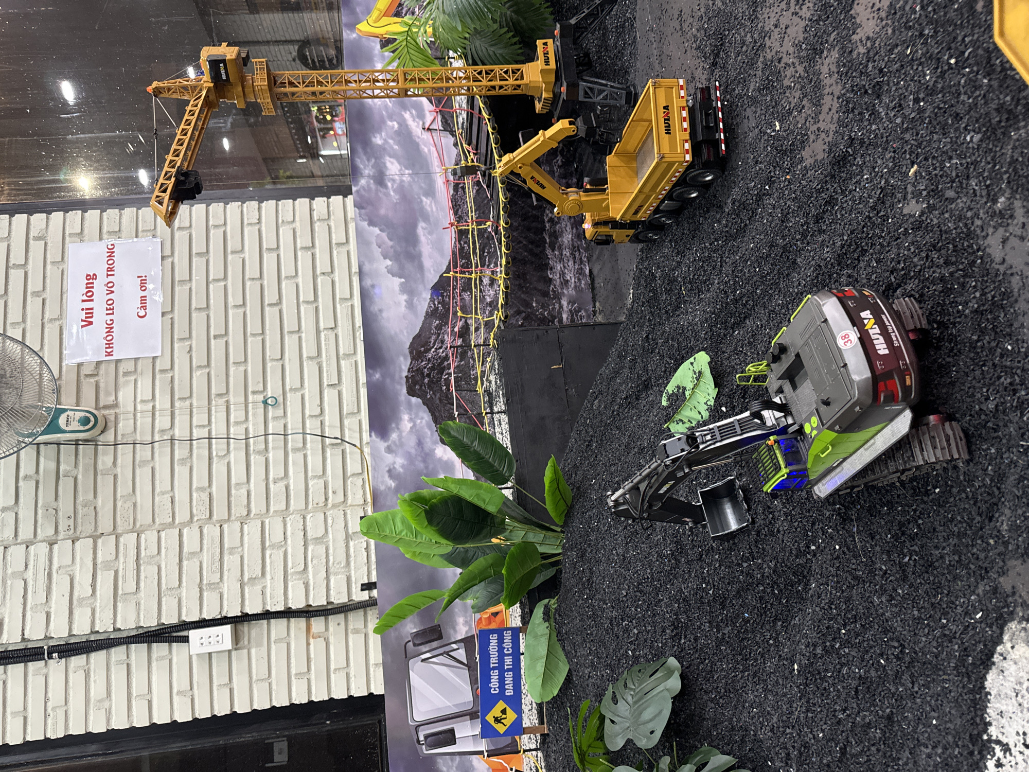 The image captures a miniature construction site exhibit, likely displayed indoors. The ground is covered with black granular material, simulating terrain. Several toy construction vehicles are arranged on this surface: a yellow tower crane is prominent on the left, a yellow dump truck with an articulating arm is in the mid-ground, and a grey and green excavator is positioned on the right. All visible vehicles are branded "HULNA." The excavator also features the text "Strong top power" and the number "38."
Artificial green plants are scattered throughout the display, and a large background poster depicts a cloudy sky over mountains, contributing to the construction site theme. A blue sign on the ground reads in Vietnamese: "CÔNG TRƯỜNG ĐANG THI CÔNG" (CONSTRUCTION SITE UNDER CONSTRUCTION).
To the left, a white brick-patterned wall is visible. A white sign with red Vietnamese text on this wall states: "Vui lòng KHÔNG LEO VÔ TRONG Cảm ơn!" (Please DO NOT CLIMB INSIDE Thank you!), indicating the display is for observation only. Above this, a white "SENKO" branded fan is mounted. An electrical outlet and black conduit are also visible on the wall. The image is rotated, with the wall appearing horizontal and the ground vertical relative to a typical upright view. The setting suggests an exhibition or public display area in Vietnam.