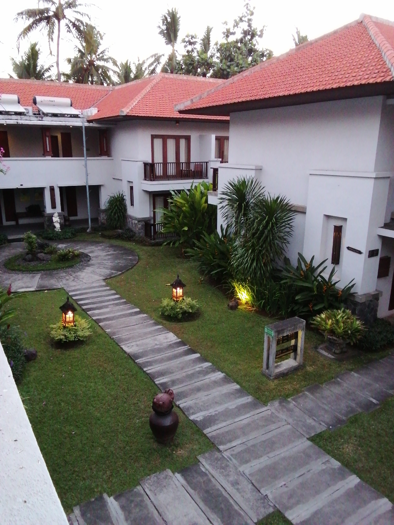 This image depicts a resort courtyard in Bulusan, Indonesia, at dusk. The scene is dominated by a white, multi-story building with traditional red tile roofing. Palm trees are visible in the background, suggesting a tropical climate. A stone pathway meanders through a well-maintained green lawn, leading towards the building. Along the pathway, two decorative lanterns are illuminated, casting a warm glow on the surrounding greenery. In the foreground, a dark, ceramic pot with a lid sits on the grass. The overall atmosphere is serene and tropical, characteristic of a vacation resort.