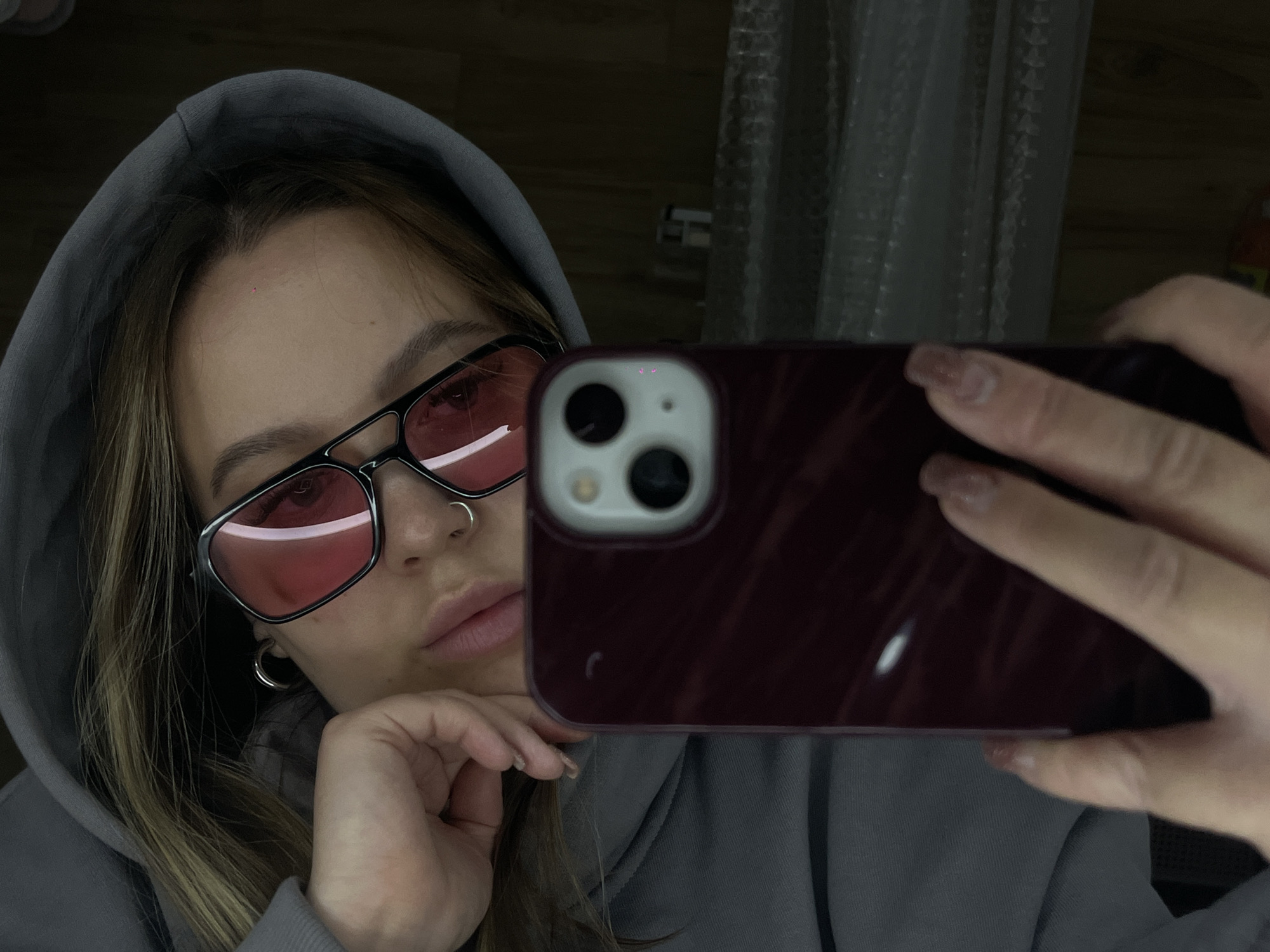 A close-up, dimly lit selfie captures a person indoors, originating from Krasnodar, Russia. The central subject, likely a young woman, wears a grey hooded sweatshirt, with light brown hair framing her face. She has on black-framed, pink-tinted sunglasses that cover her eyes, reflecting a faint light source. A silver nose ring is visible on her septum, and a small silver hoop earring adorns her left ear. Her lips are slightly parted in a neutral expression.
She is holding a dark red or maroon smartphone, possibly an iPhone, with her right hand, positioned to take the selfie. The phone's multi-lens camera module is prominently visible in the reflection. Her nails are long and appear to have a light, possibly nude, polish. The background is dark and out of focus, showing only vague shapes, including what might be a curtain or textured surface to the right. The overall mood is casual and slightly introspective due to the low light and the person's expression. No discernible text is visible in the image.