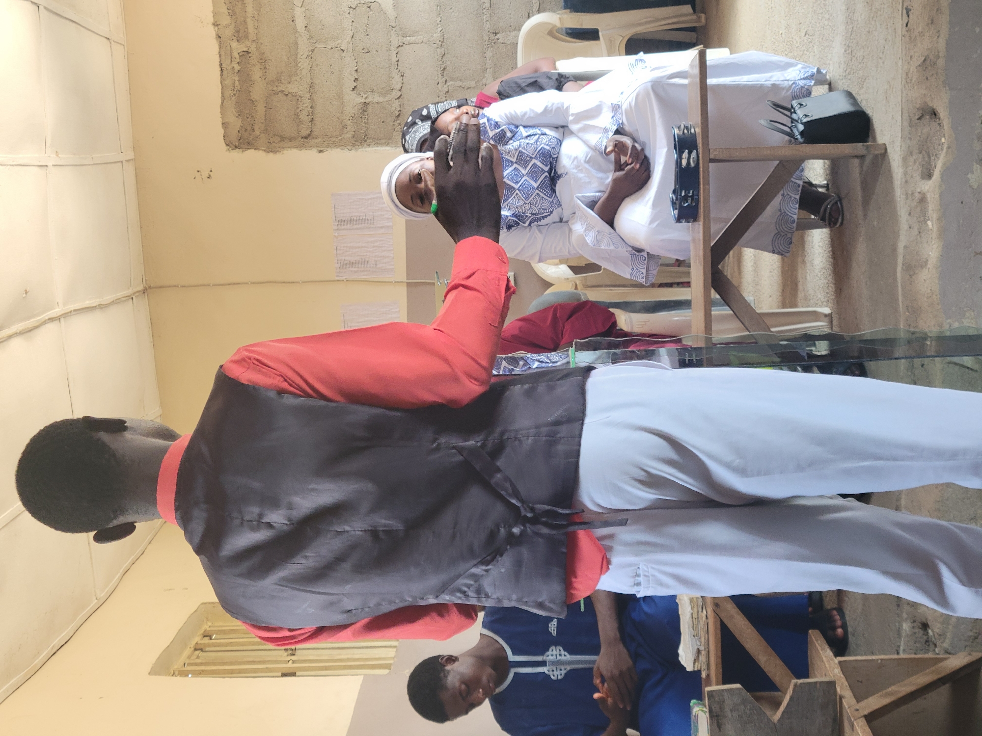 In a room in Madalla, Nigeria, a man in a red shirt and black vest stands with his back to the camera, holding a hand near the face of a woman in a white dress and headscarf. The woman is seated, facing the man, with a pattern of blue on her dress. Behind her, another woman with a patterned headscarf is also seated. To the left, a young man in a blue and white robe sits with his hands clasped, looking down. He is seated at a wooden table with books on it. A wooden chair is visible behind him. The room has cream-colored walls, a drop ceiling, and a concrete block wall section on the right. A black handbag rests on the floor next to a wooden table in front of the seated women, where a tambourine is also placed.