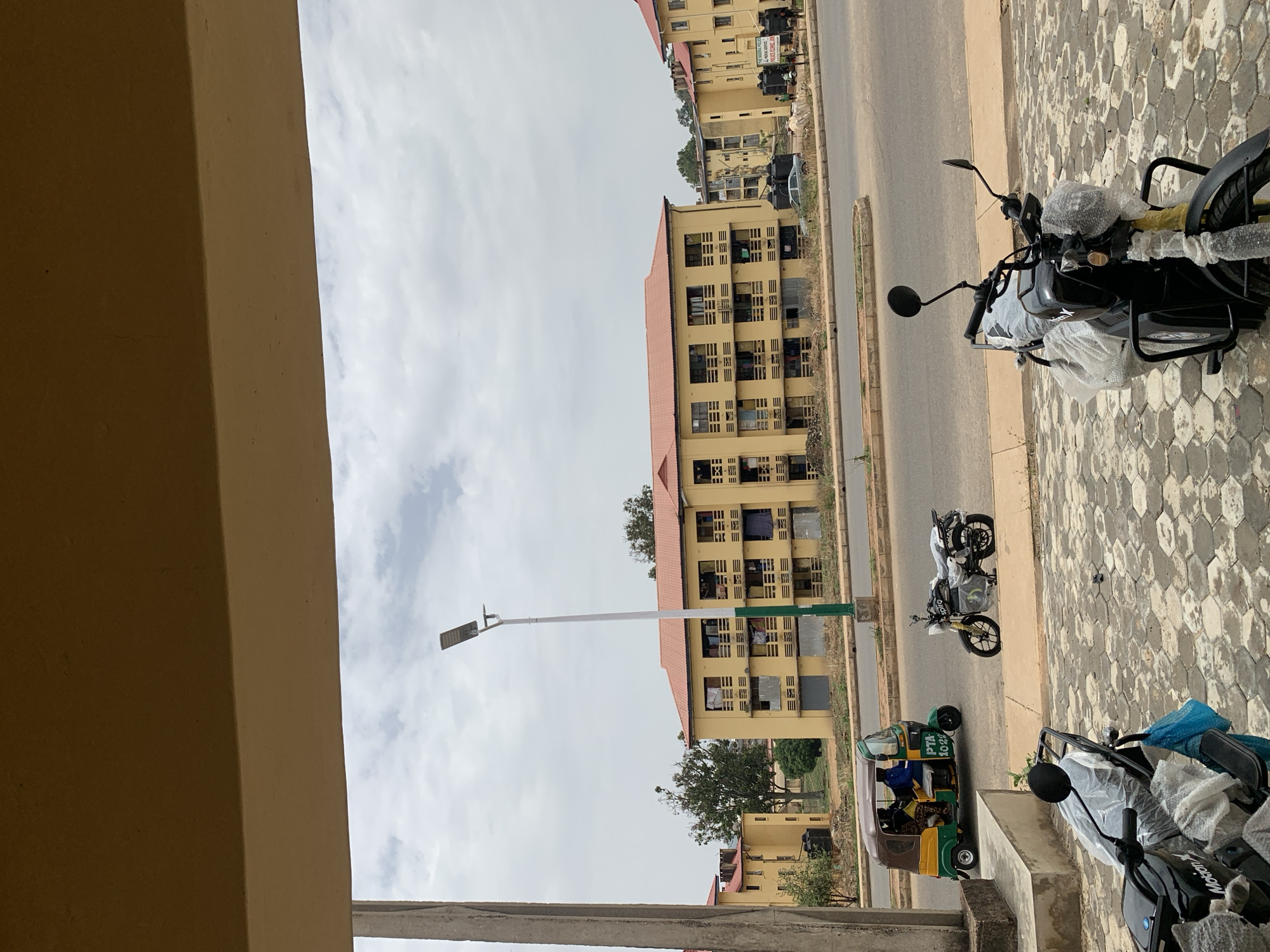The image shows a street scene in Jos, Nigeria. Several multi-story, yellow buildings with red roofs line the street. A light pole stands near one of the buildings. The road has a green and yellow tricycle taxi and two motorcycles on it. Two more motorcycles are parked on a paved area with a hexagonal pattern. The sky is overcast. The viewpoint is from an elevated position, partially obscured by a beige-colored architectural feature.