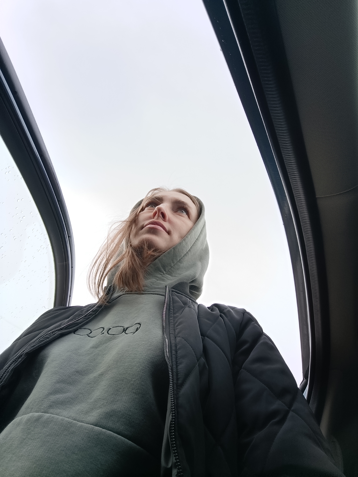 A low-angle shot shows a young woman indoors, seemingly looking upwards. She is wearing a dark, quilted jacket over a green hoodie. Water droplets are visible on what appears to be a glass window or panel to the left. The background includes parts of the interior structure, possibly a vehicle sunroof, and an overcast sky.
While the location within Chaikovsky, Russia, cannot be definitively determined from the visual information, the overall setting suggests an enclosed space with access to the outdoors, such as a car or building with a glass-enclosed area.