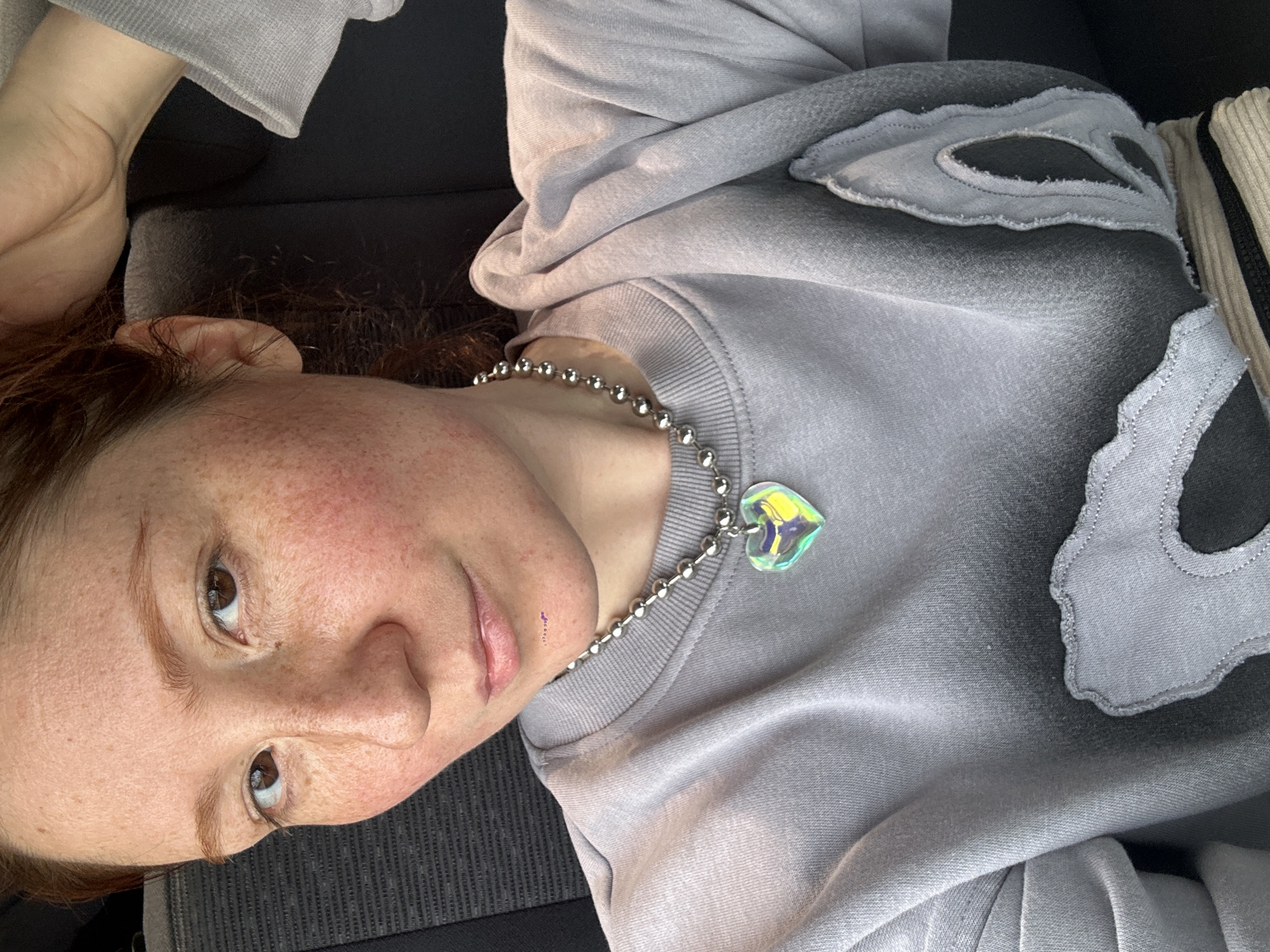 A pale-skinned person with reddish-brown hair, freckles, and brown eyes is lying on a dark surface, possibly a car seat, wearing a gray sweatshirt and a silver ball-chain necklace with a heart-shaped pendant. The sweatshirt has decorative gray patches. Part of a cream-colored bag with a black zipper is visible. The location is unknown and cannot be determined from the image.