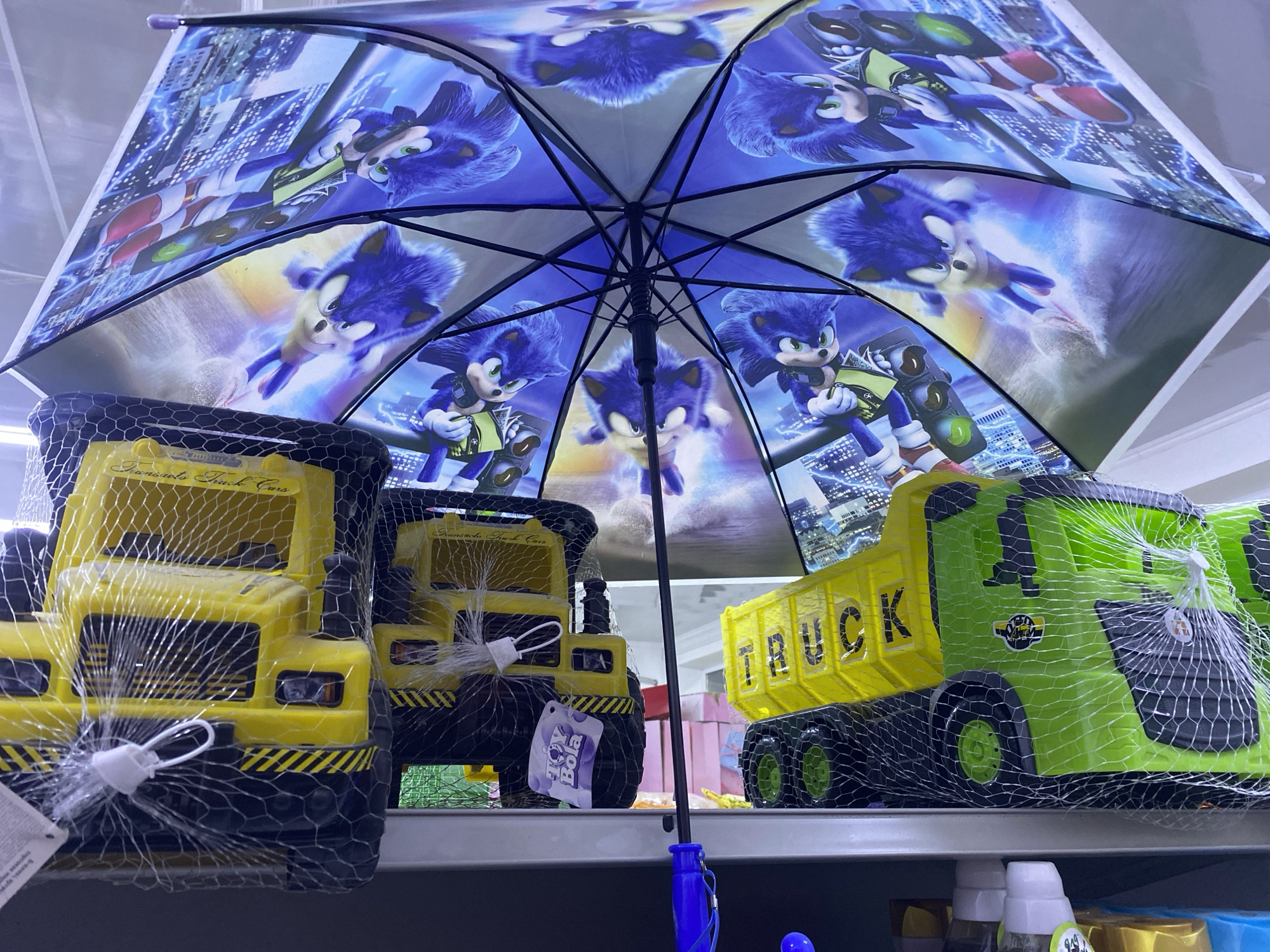 The image shows a close-up of an open umbrella with a Sonic the Hedgehog theme. The umbrella is decorated with multiple images of Sonic in various action poses, set against a backdrop of cityscapes and lightning effects. Below the umbrella, two large toy trucks are visible on a shelf. The truck on the left is yellow and has "Fremonts Truck Cars" printed on its side. The truck on the right is yellow and green and has "TRUCK" printed on its side. Both toy trucks are wrapped in a mesh netting. The setting appears to be a retail store, likely a toy section. There are no discernible cues about the time of day or weather. The emotional tone is neutral, focusing on the presentation of the merchandise.