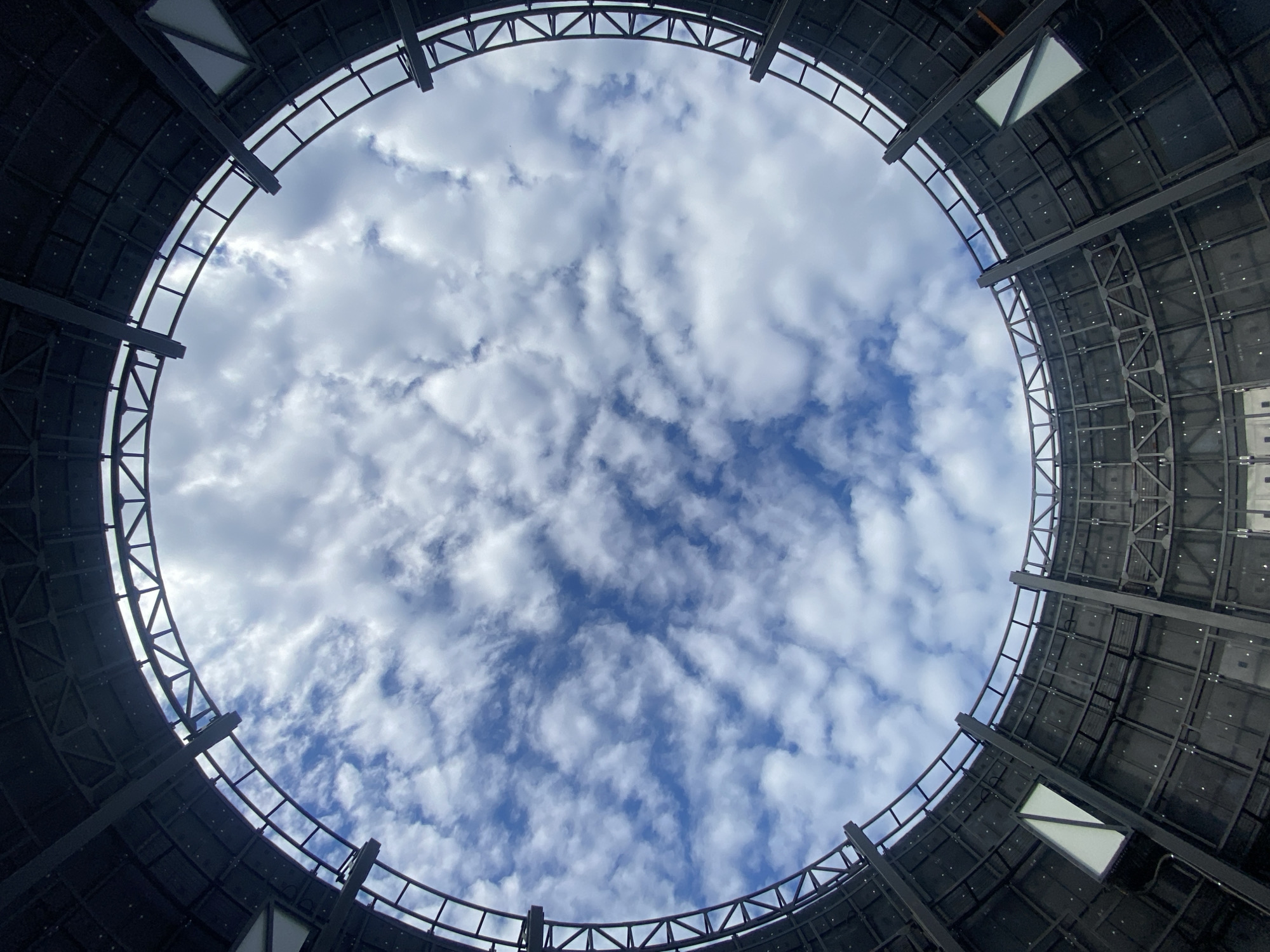 Looking up from inside a large, cylindrical structure, a circular opening reveals a sky filled with white, puffy clouds against a backdrop of blue sky. The structure’s interior is constructed of metal girders and panels, with small, rectangular openings interspersed along the walls. The location is Заволжье, Russia.