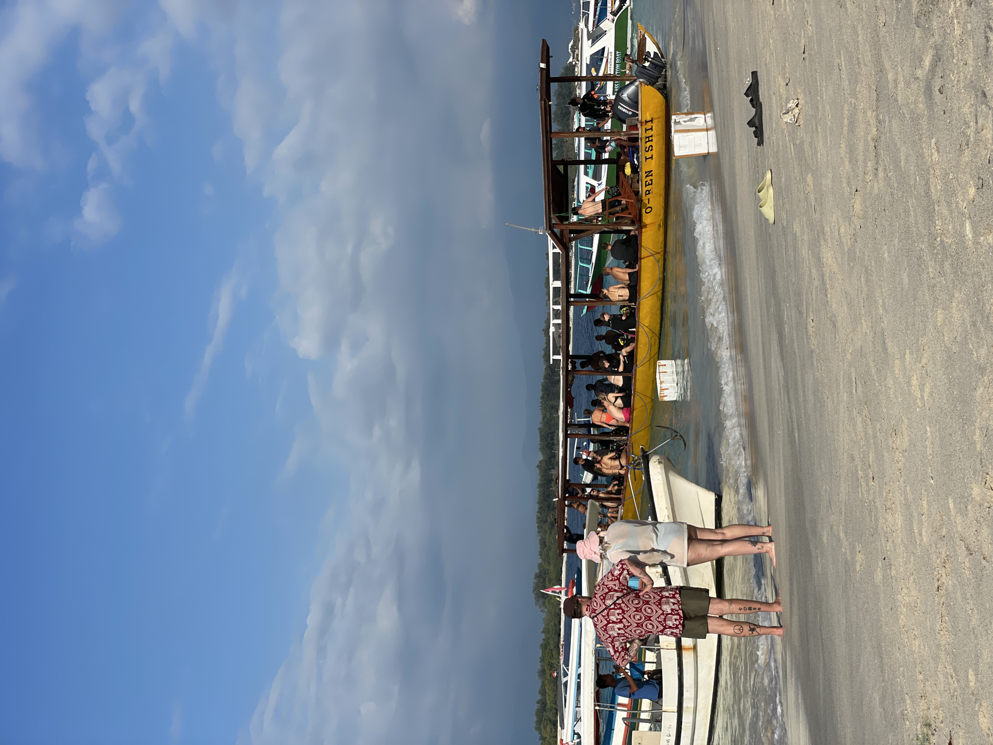 In Unknown, Indonesia, at a beach, two individuals stand on the sand near the water's edge. The person on the left, wearing a red shirt with a white pattern and khaki shorts, faces away from the camera. The person on the right, in a white sheer dress and a pink hat, also faces away. Their legs display tattoos.
Several boats are present in the shallow water. A large yellow boat, named "O-REN ISHII," is filled with passengers. Another white boat is docked to its left, with a person in a blue shirt standing on it. In the background, more boats are visible, including one with green accents. The sky is blue with scattered clouds. The environment appears to be a tropical beach with sand, clear water, and distant green land.