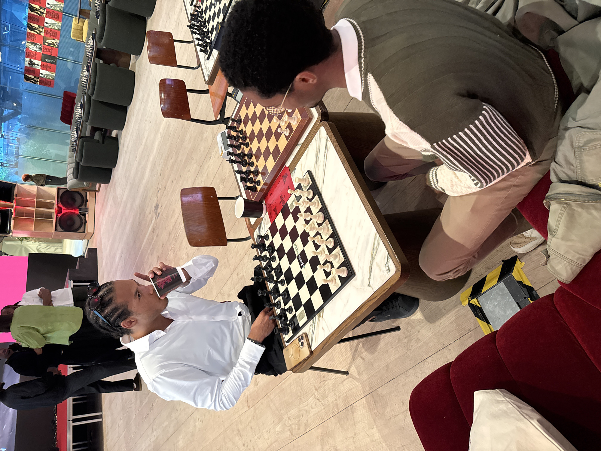 The image captures an indoor scene in what appears to be a lounge or recreational area, possibly in London, United Kingdom, given the context. Two African American males are deeply engrossed in a game of chess. The male on the left side of the image is dressed in a white collared shirt. He is holding a cup. His opponent is wearing a green and white sweater. There are three chessboard tables arranged in a row with several chess pieces on them. Numerous black and green chairs can be seen in the background. A display on the wall depicts images of various subjects. A red velvet couch is on the right.