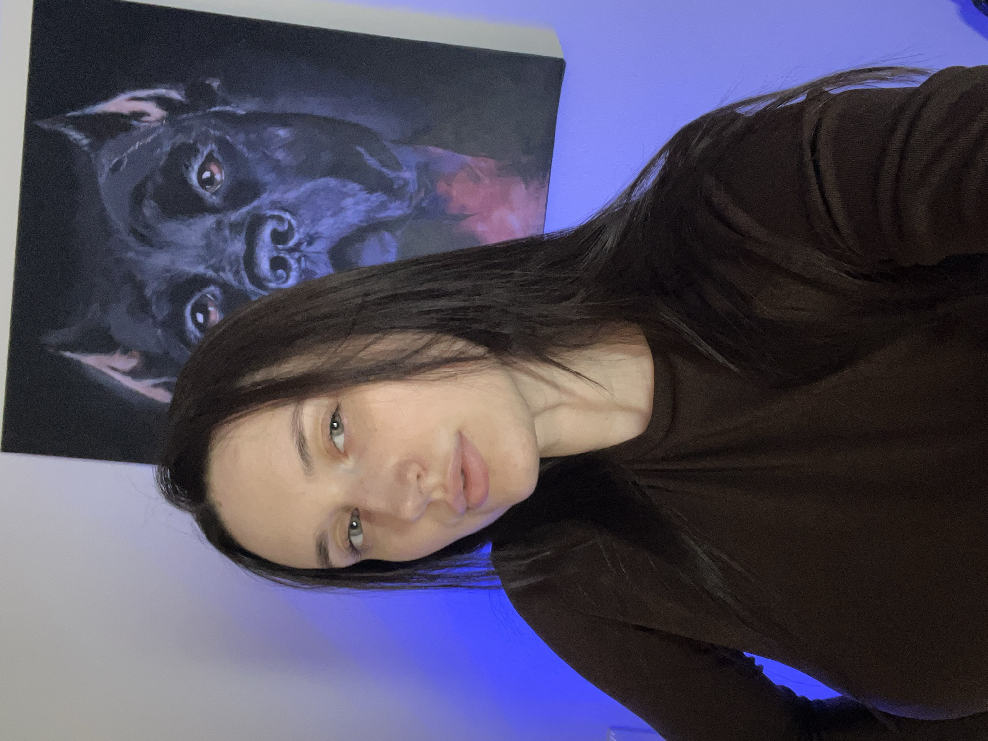 A woman with long, dark hair and light skin poses in front of a painting of a black dog. She is wearing a brown long-sleeved shirt. The painting of the dog is mostly black and appears to be a close-up of the dog's face. The background is a wall with a blue glow. There is nothing to indicate that this is in Tyumen, Russia.