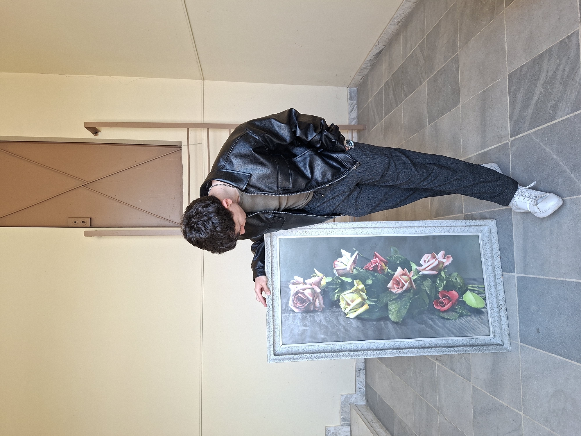 A young man in a black leather jacket and grey trousers stands on a tiled floor in Plovdiv, Bulgaria, holding a framed picture of roses. He is looking at the picture, which is resting on the floor. The background consists of cream-colored walls and a brown door. The setting appears to be an interior hallway.