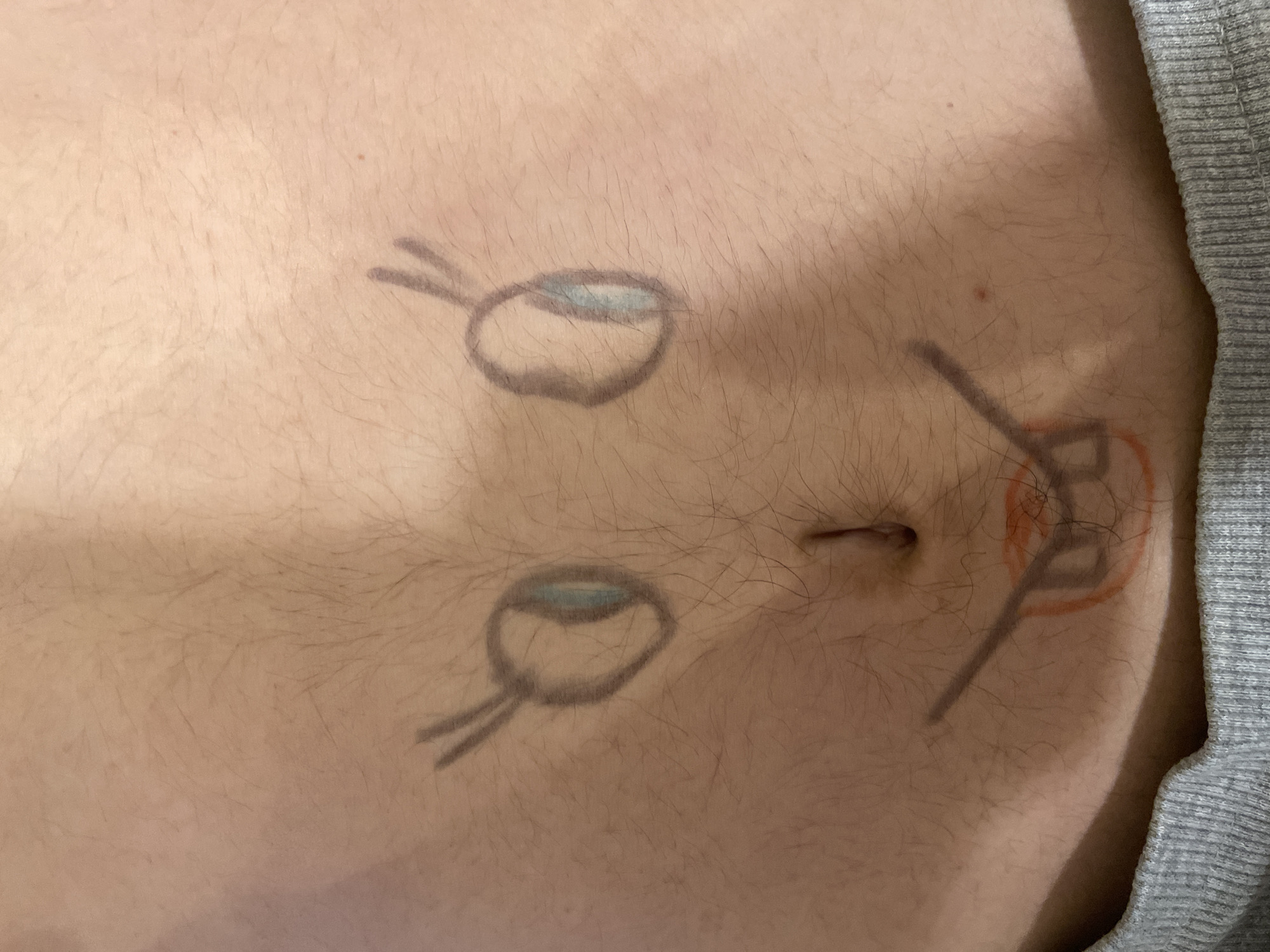 A close-up shot shows a torso with a belly button. Two oval shapes resembling eyes with short lines extending from one side are drawn above the belly button. To the right of the belly button, near the edge of the image, there is another drawing featuring straight lines and a partial orange circle surrounding it. The skin is light-colored and has body hair. A portion of a gray, textured garment is visible along the right edge of the frame.