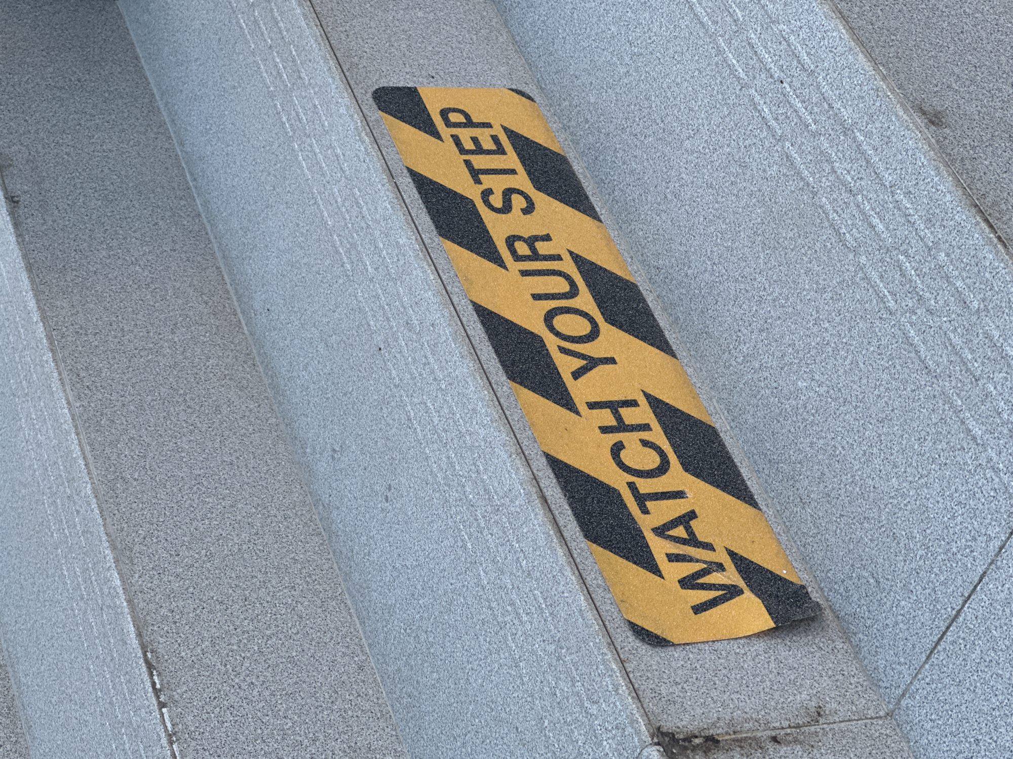 A high-angle, close-up shot captures a section of outdoor stairs in Budva, Montenegro, constructed from light grey, granular material, likely granite or speckled concrete. A prominent yellow and black anti-slip strip is affixed diagonally across one of the steps, serving as a safety measure. The text "WATCH YOUR STEP" is clearly visible in black capital letters on the yellow sections of the strip, oriented to be easily read by individuals using the stairs.
The surface of the stairs features subtle etched lines or grooves, particularly on the risers and the edges of the treads, which may provide additional traction or be decorative. The lighting is bright and natural, suggesting it is daytime with clear weather conditions. While no people or specific activities are depicted, the presence of the safety strip implies that these stairs are in regular use and that caution is advised for pedestrians. The texture of both the stair material and the rough, sandpaper-like anti-slip strip is discernible, emphasizing their practical function.