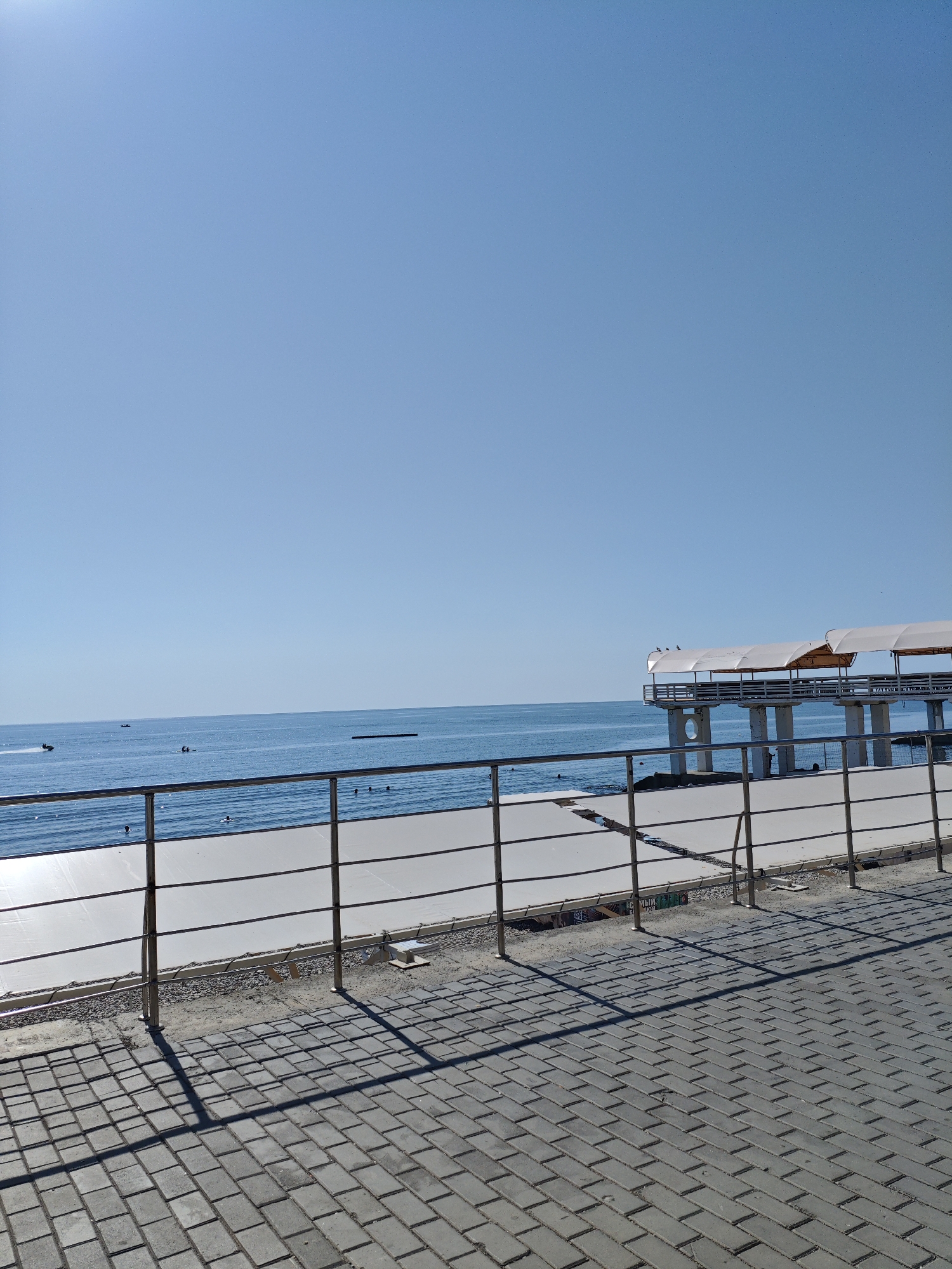 This is an eye-level, medium shot photograph of a seaside promenade in Alushta, Ukraine. The sky is a clear, bright blue. In the foreground, a paved walkway made of grey interlocking bricks is visible, with long shadows cast by a metal railing. The railing, made of thin metal poles and horizontal bars, runs along the edge of the promenade. Beyond the railing, there is a strip of gravel and then a white, flat surface that appears to be under construction or being cleaned. Further beyond this surface, the blue sea stretches out to the horizon, with a few small boats and people visible in the water. On the right side of the frame, a pier extends out over the water, supported by concrete pillars. The pier has white canopy structures overhead and a few birds are perched on them. The overall scene depicts a calm, sunny day at the coast.
