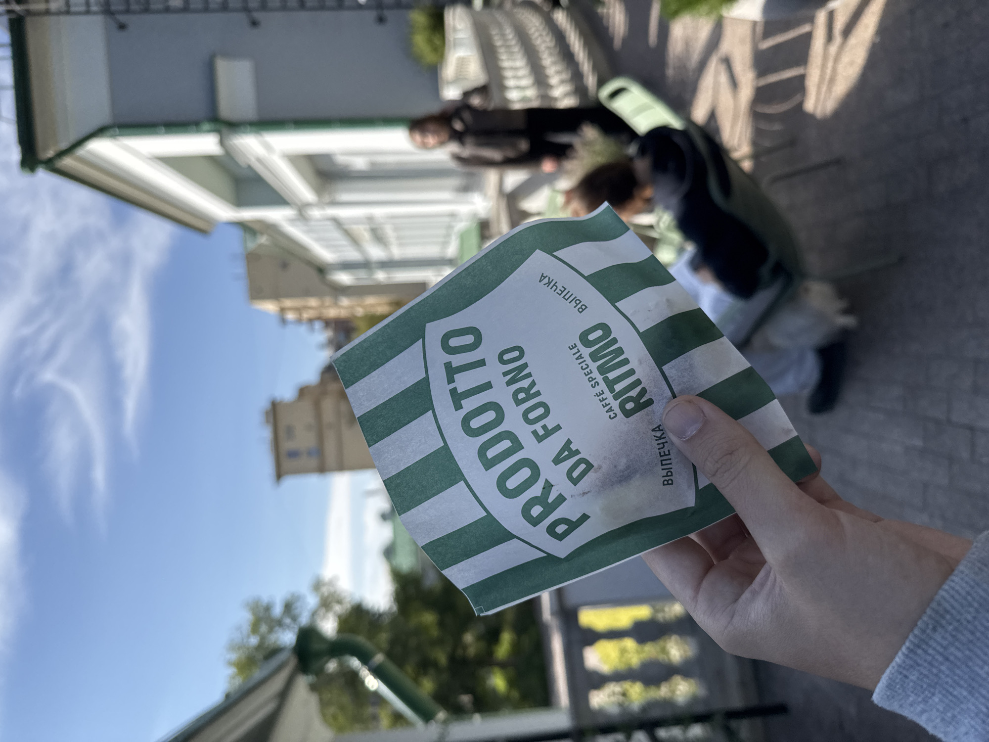 A hand holds a green and white striped paper bag with the words "PRODOTTO DA FORNO" printed on it. Below this, in smaller text, are the words "CAFFÉ SPECIALE RITMO" and "ВЫПЕЧКА". The background shows a bright, sunny day with a blue sky and some clouds. In the background, there is a building with white trim and balconies, and a person in a dark jacket standing on a balcony. Another person is seated in a green chair in the foreground. The image appears to be taken outdoors on a terrace or courtyard.