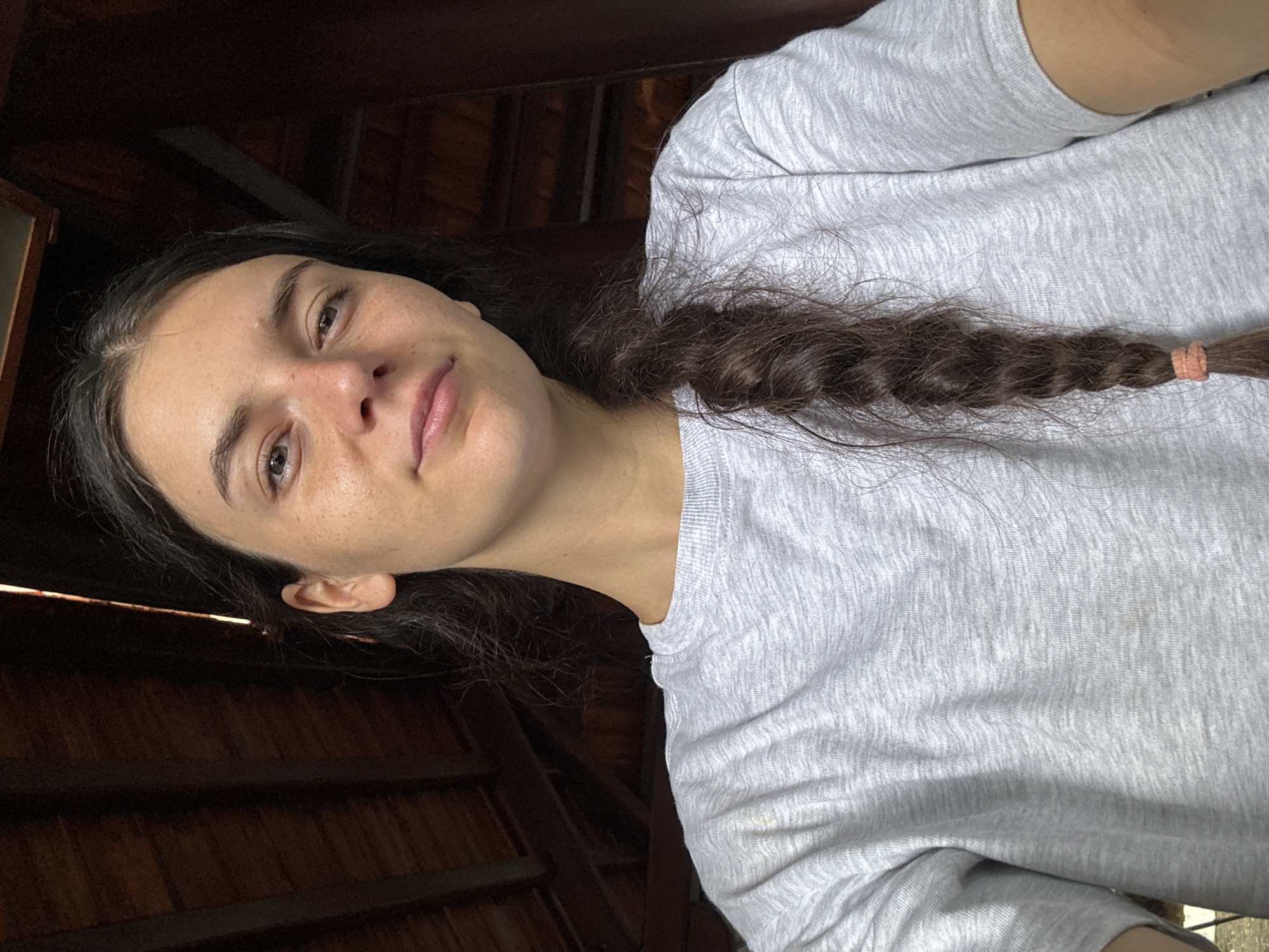 The image is a portrait-style selfie of a young woman, captured from a slightly low angle and oriented vertically with her head tilted to the left. The woman has dark, possibly brown, long hair pulled back from her face and styled into a thick braid that rests over her right shoulder, secured with a peachy-pink elastic band. She has natural-looking skin with visible freckles on her nose and cheeks, and brown eyes. Her expression is calm and relaxed, with a slight, subtle smile. She is wearing a light grey, long-sleeved t-shirt with a textured fabric, which shows a faint yellowish spot on the lower right side.
The background consists of dark wooden slatted panels, suggesting an indoor or covered outdoor setting, possibly a hut or a shaded area characteristic of tropical locations. A narrow strip of light is visible higher up in the background, indicating an opening or a source of natural light from outside. The overall lighting is somewhat dim but even, casting soft shadows. The setting, combined with the location context of Mũi Né, Vietnam, suggests a warm, possibly rustic or resort-like environment. No visible text is present in the image.