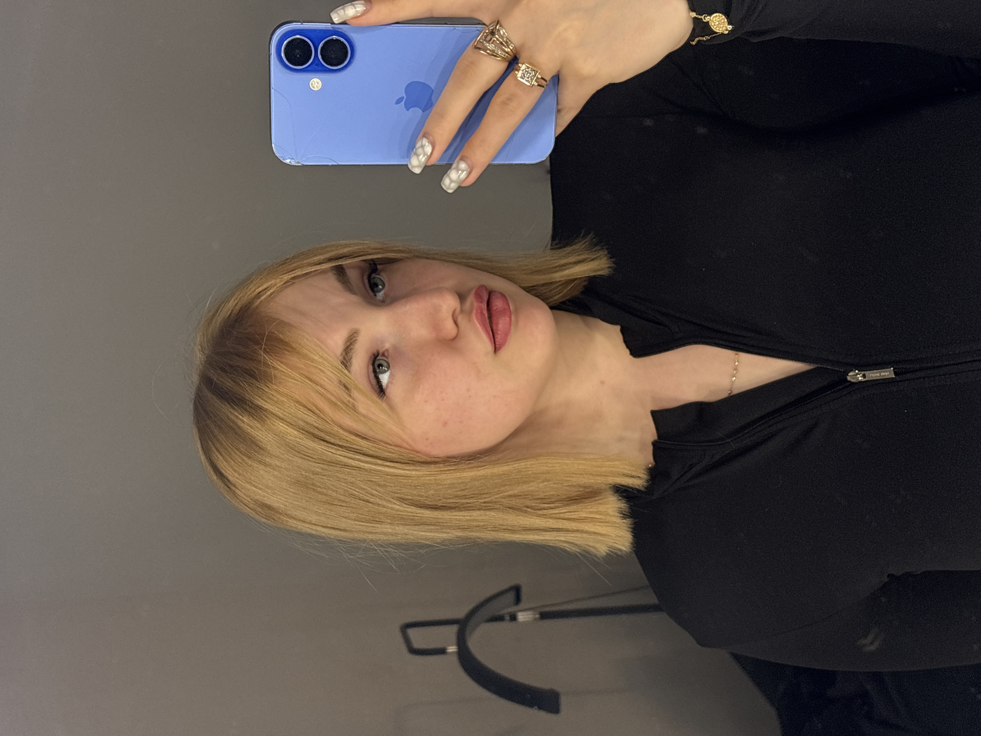 A young woman with blonde hair and blue eyes is taking a selfie in a room. She is wearing a black zip-up shirt and several rings. She is holding a blue iPhone in her right hand, which is angled to capture her reflection in a mirror. The background is a plain gray wall, with a black floor lamp visible behind her on the left. The overall scene is simple and uncluttered, suggesting a personal space. The image does not provide any information about the city of Khimki, Russia.