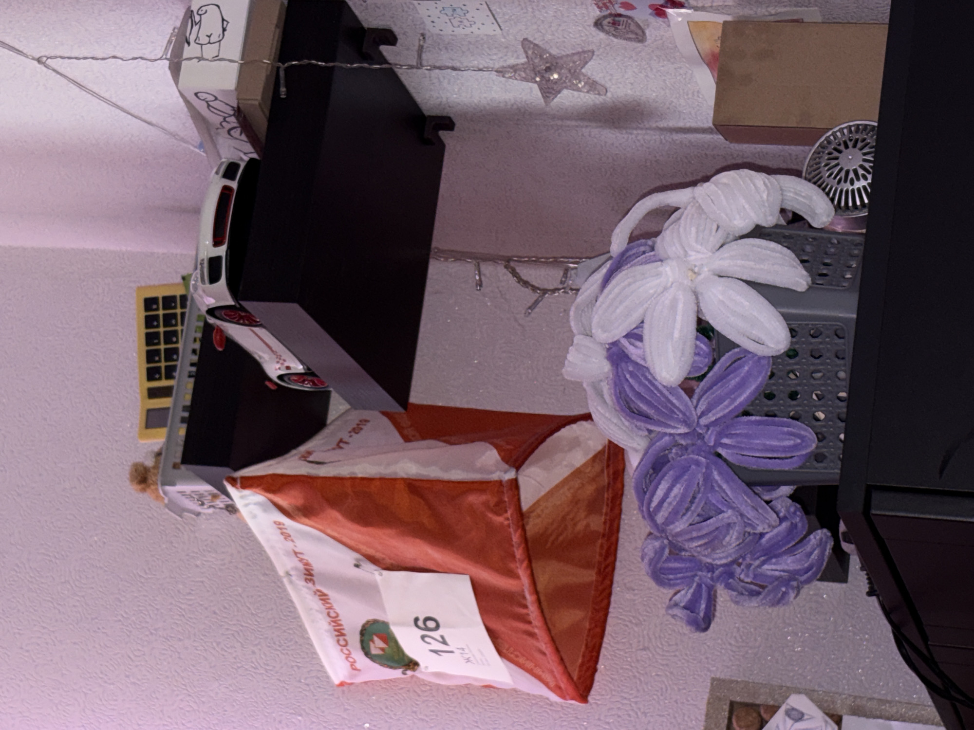 The image depicts a room interior in Прикамский, Russia. Dominating the view is an orienteering flag with the number "126" and Russian text "РОССИЙСКИЙ ЗИЮТ-2019". Adjacent to the flag are two floral decorations: one white and one purple. These sit near a dark-colored shelving unit or table. Various other objects are scattered around, including a white box with cartoon drawings, a toy car, a calculator, a star-shaped decoration, a cardboard box, and a metal drain cover. The environment is enclosed, with a textured wall serving as the backdrop.