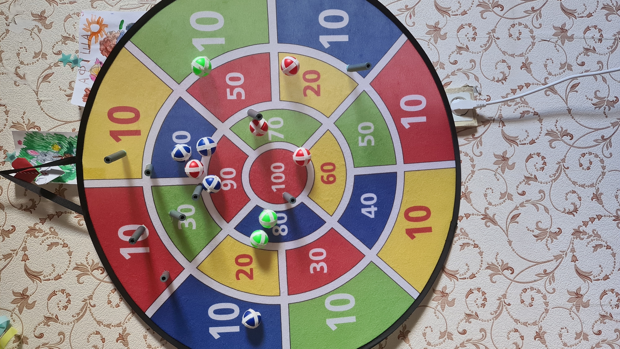 A colorful dartboard hangs on a wall with a patterned wallpaper. The dartboard is divided into sections with numbers and colors, including red, blue, yellow, and green. Several darts are stuck in the board, some in the numbered sections and some in the bullseye. There are also some darts that have landed near the board. Two children's drawings are attached to the wall above the dartboard. A power outlet with a white cord plugged into it is visible on the wall below the dartboard. The scene appears to be indoors.