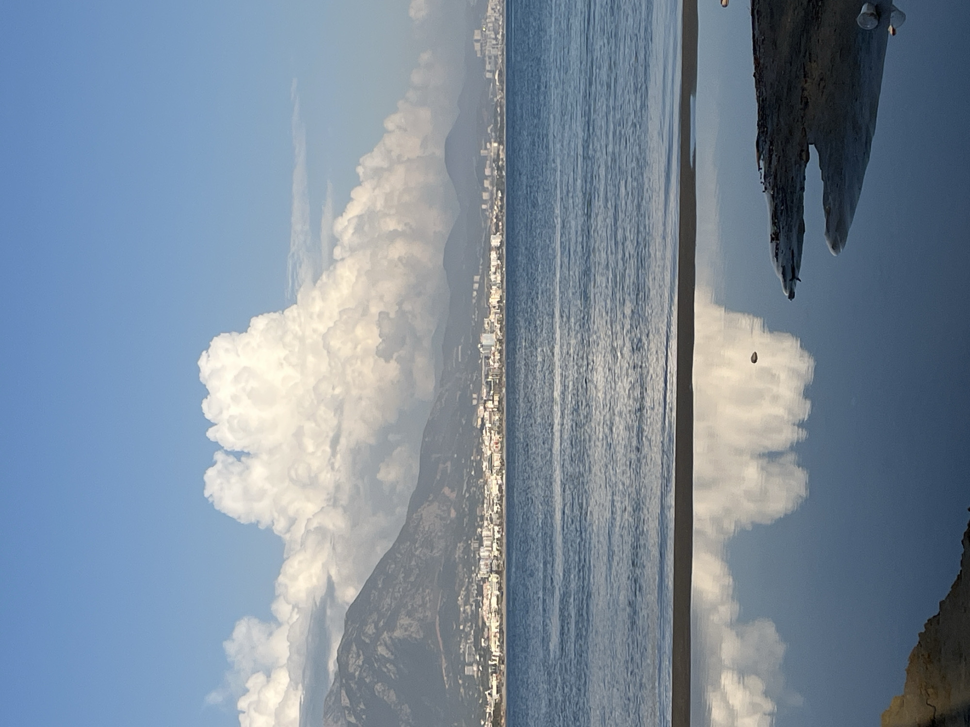 This image depicts a coastal scene in Alanya, Turkey. On the left is a large, cumulonimbus cloud formation dominating the sky behind a mountain range. A strip of buildings, presumably the city of Alanya, stretches along the base of the mountain and along the coastline. The right portion of the image shows the surface of the sea, with reflections of the sky and clouds visible on the water. In the foreground, there is a rocky outcrop with an eroded shape jutting out into the sea. The water is calm with a slight ripple.