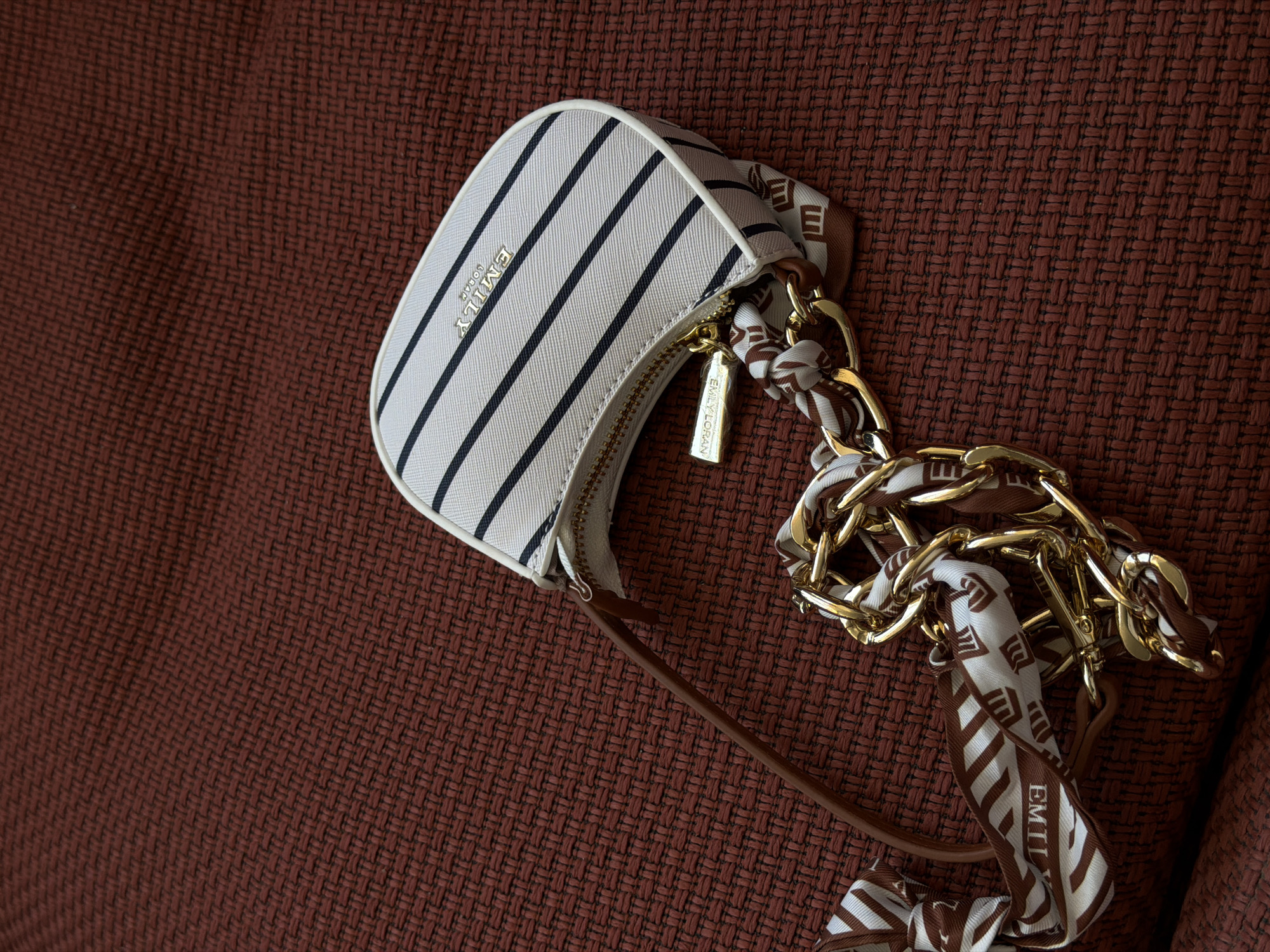 The image shows a small, striped handbag placed on a textured brown surface. The bag is primarily white with black vertical stripes, and the brand name "EMILY LONDON" is visible in gold lettering on the front. The bag has a gold zipper and a brown shoulder strap. A gold chain with a patterned scarf woven through it is attached to the bag. The background appears to be a woven fabric, possibly a blanket or upholstery. The location is unspecified in the image, and there is no information to indicate this scene is in Bishkek City, Kyrgyzstan.