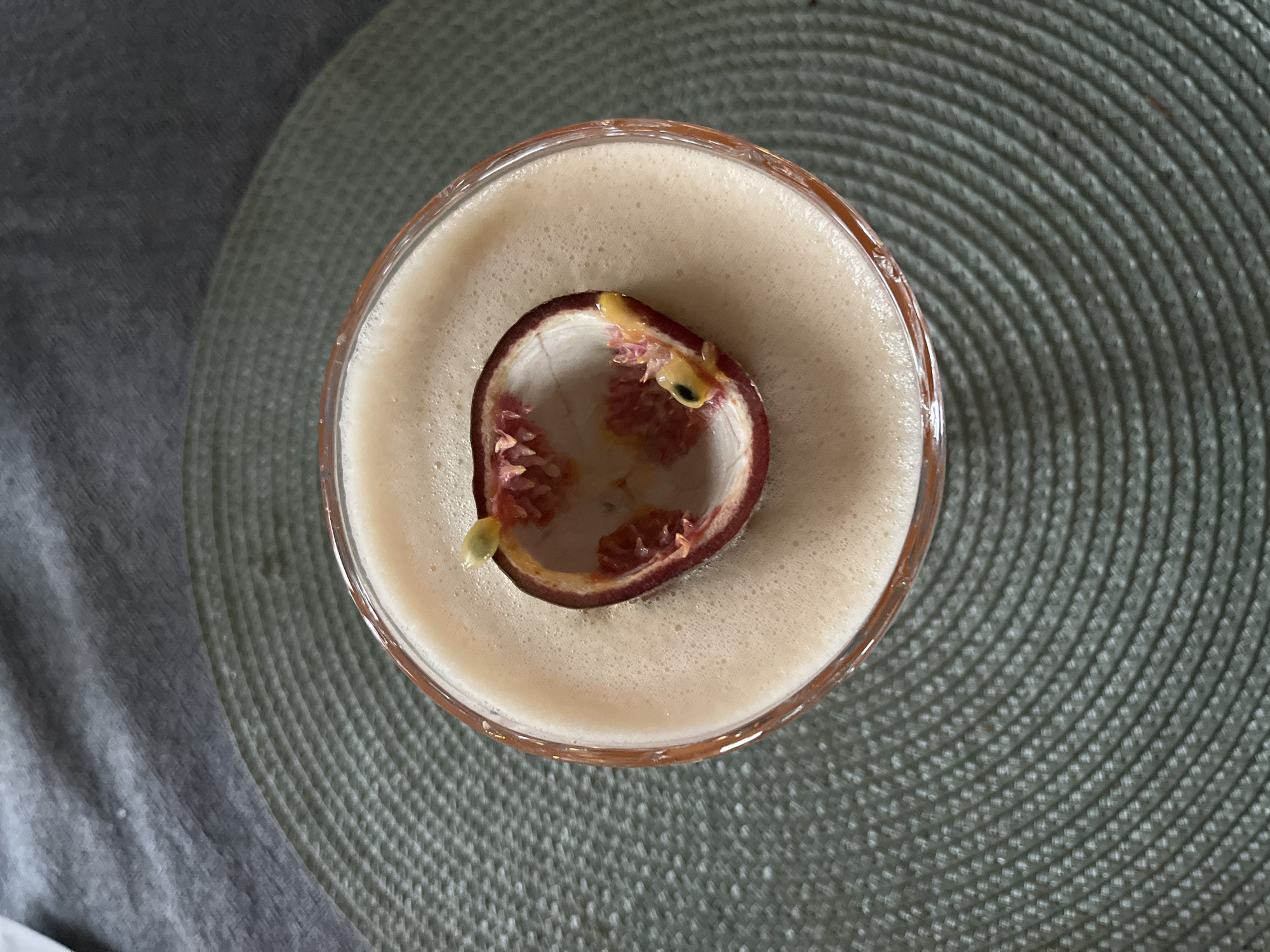A close-up, overhead shot captures a cocktail in a glass. The drink has a frothy, light pinkish-beige surface, and a piece of passion fruit peel, containing some seeds, floats on top. The glass is situated on a round, textured green placemat. In the background, a grey fabric surface is visible. The image appears to be taken indoors, likely in a domestic setting. There is no discernible information about the location in Kongsberg, Norway.