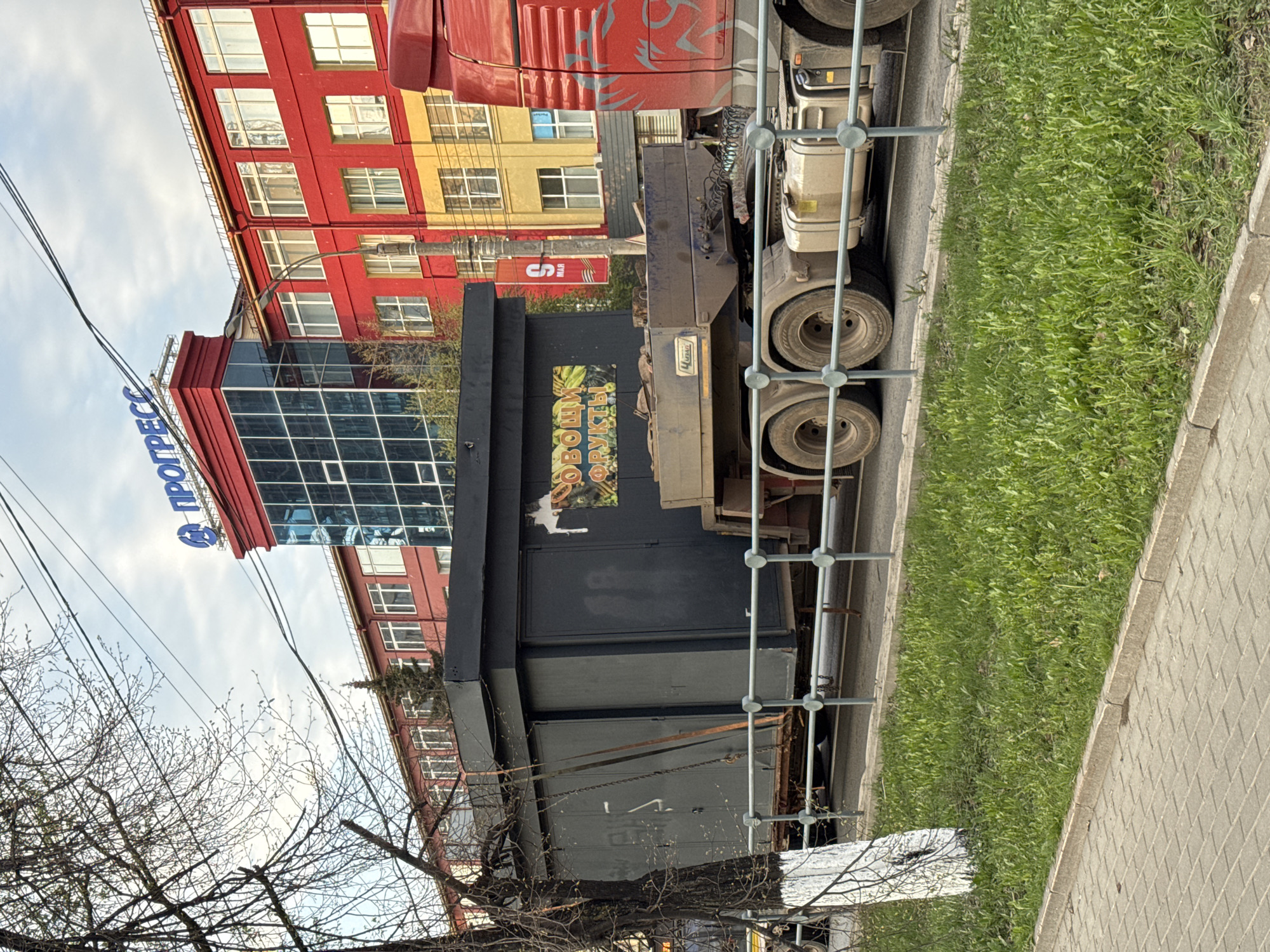 The image depicts a street scene in what is presumed to be Izhevsk, Russia, based on the Cyrillic script on the "Progress" building signage. A large truck, possibly a delivery vehicle, is parked beside the sidewalk. Adjacent to the truck is a green, grassy area. The background consists of a multi-story building with a combination of red and yellow exterior walls and glass windows. A smaller, glass-fronted building is attached to the taller structure, bearing the "Progress" sign. Bare tree branches are visible in the lower-left corner of the image. Overhead utility lines stretch across the sky.