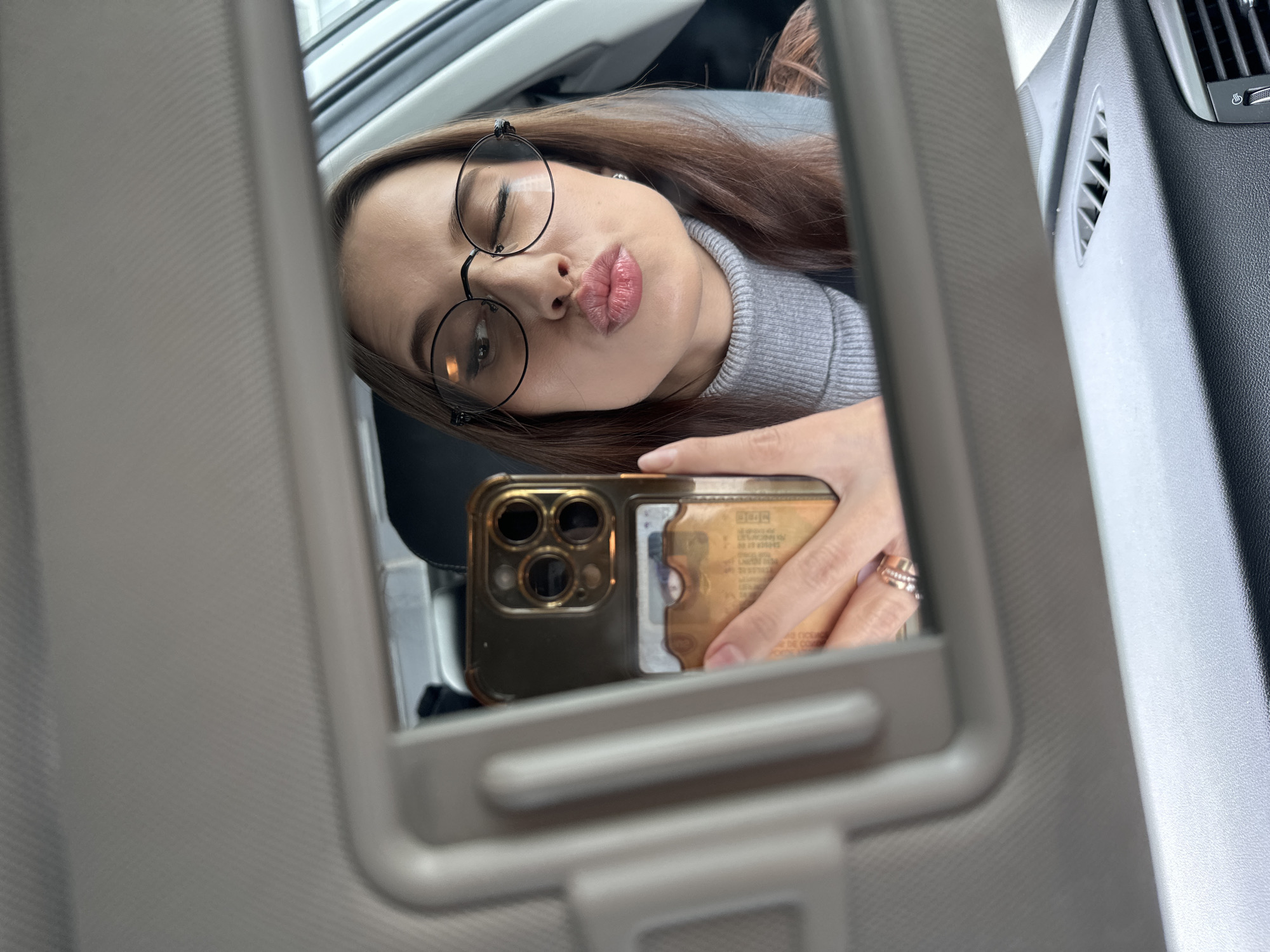A woman is captured in a selfie taken through a car's vanity mirror, reflecting her image. She has long brown hair and is wearing round glasses. Her eyes are closed, and she is making a playful "pout" with her lips. She wears a grey turtleneck sweater.
She is holding a smartphone, likely an iPhone with three camera lenses, encased in a clear or gold-toned case. A card, possibly an ID or credit card, is visible tucked into the back of her phone case, though its text is blurred and illegible. A metallic ring, possibly a stacking ring set or a wide band, is visible on her ring finger.
The setting is the interior of a car, specifically using the passenger-side sun visor mirror for the reflection. Parts of the car's dashboard and air vents are visible on the right side of the frame, indicating a modern vehicle interior. The lighting suggests it is daytime. There are no specific visual cues within the image itself to confirm the stated location of Yekaterinburg, Russia.