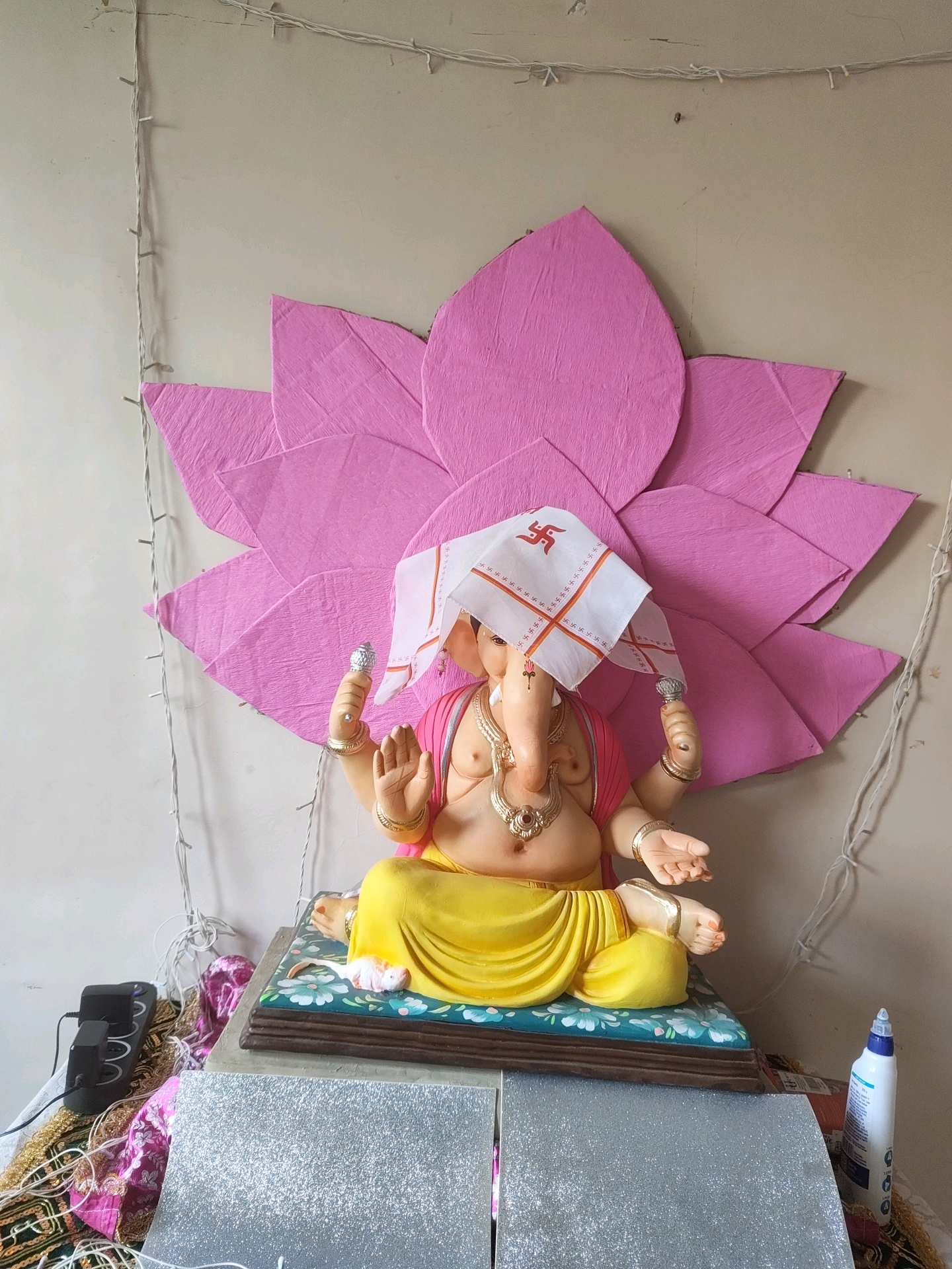 The image shows a statue of Lord Ganesha, a prominent Hindu deity, adorned with a pink top and a yellow dhoti. A white cloth with a red Swastika symbol is draped over its head. The statue is placed on a decorative base and is set against a backdrop of large, pink, lotus-shaped paper cutouts. String lights are visible in the background, draped along a light-colored wall. To the left of the statue, a black power strip with multiple outlets is placed on a colorful fabric. To the right, a white bottle with a blue cap is standing. The overall scene appears to be a home altar or decoration, likely for a religious festival. The image does not provide any specific information about the city of Pune, India, or any details about people or actions.