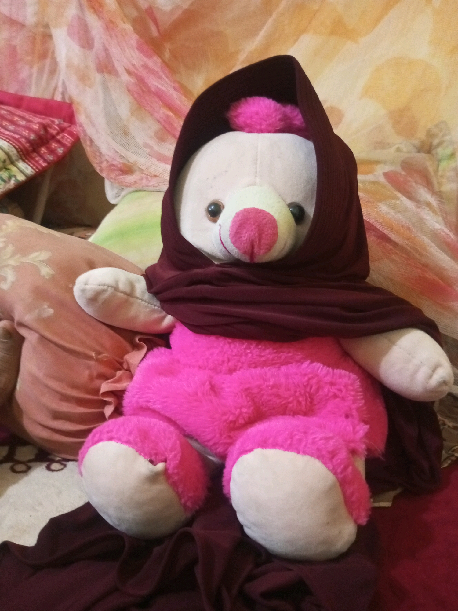 A bright pink teddy bear with a white face, nose, and paws is sitting upright on a bed. The teddy bear is wearing a dark maroon headscarf that covers its ears and is draped around its neck and shoulders. The teddy bear has black button eyes and a pink nose. It is positioned in the center of the frame, with pillows and patterned fabrics in the background, suggesting an indoor setting likely in a bedroom in the city of Bundar, Indonesia.
