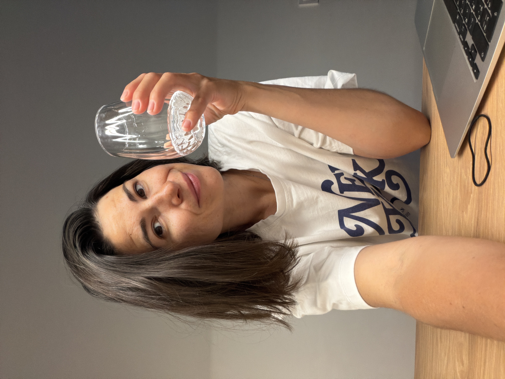 A person is seen holding a clear glass. The person is wearing a white t-shirt with a dark blue design. A laptop is on a wooden desk. The background is a gray wall. The location is not discernable.