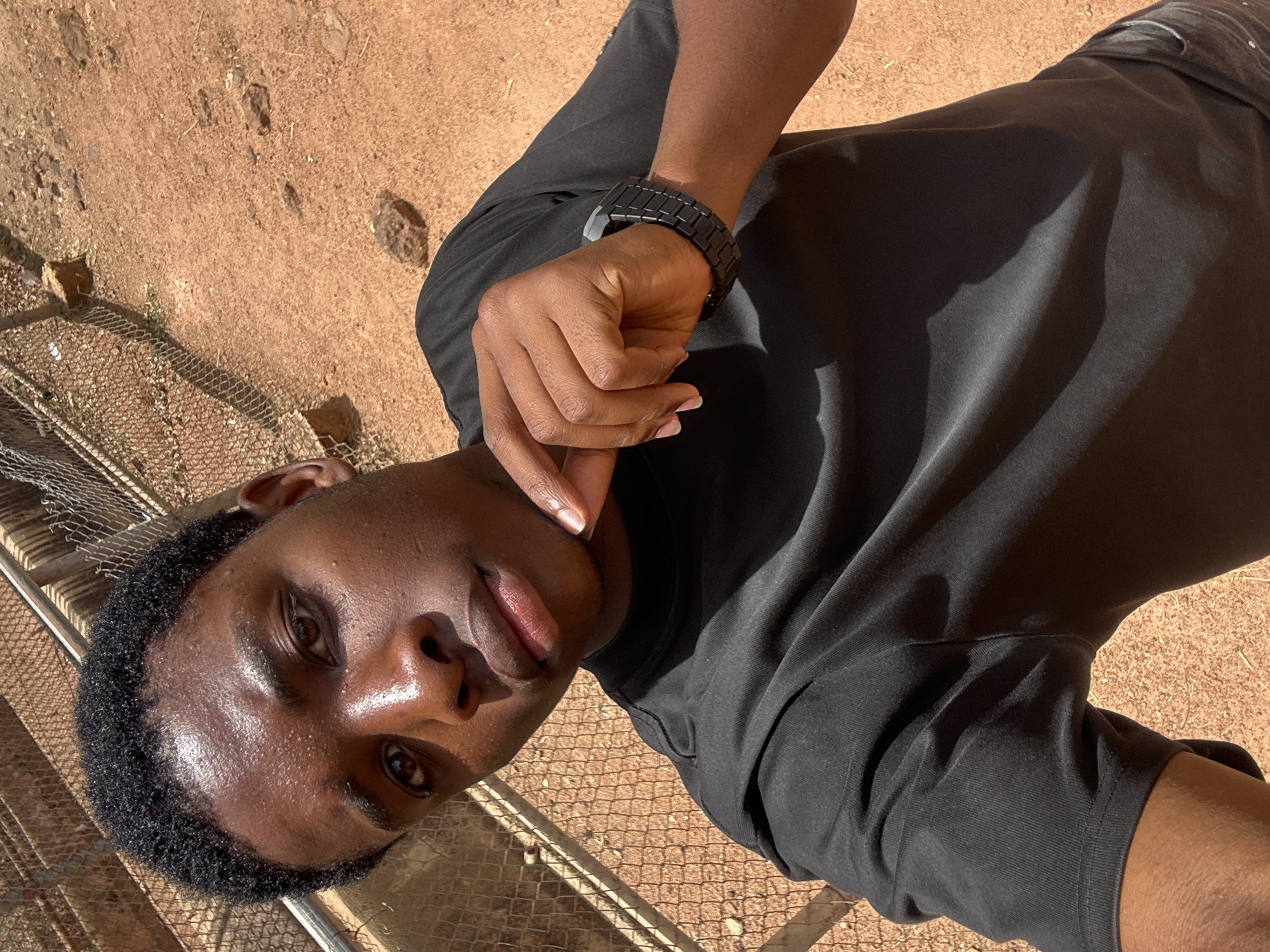 A young Black man is depicted in a close-up, high-angle portrait, captured outdoors under bright, direct sunlight. His head is tilted to the side as he looks directly at the camera with a relaxed expression, featuring a slight smirk with his tongue subtly visible between his lips. His short, curly black hair frames his face, which shows natural sheen, possibly from sweat or oil, highlighting his forehead, nose, and cheekbones. His dark brown eyes are clearly visible. His right index finger is gently resting near his chin and lower lip.
He is wearing a plain black short-sleeved t-shirt. On his left wrist, a sleek, black digital watch with a segmented metal band is visible. The background primarily consists of reddish-brown, sandy or dusty ground, typical of a dry, outdoor setting, consistent with the landscape in Jos, Nigeria. A metal mesh fence or grating can be seen in the lower left portion of the image and extending behind his head. The strong sunlight casts distinct shadows, indicating a clear, sunny day, likely during mid-day or early afternoon. No visible text is present in the image.