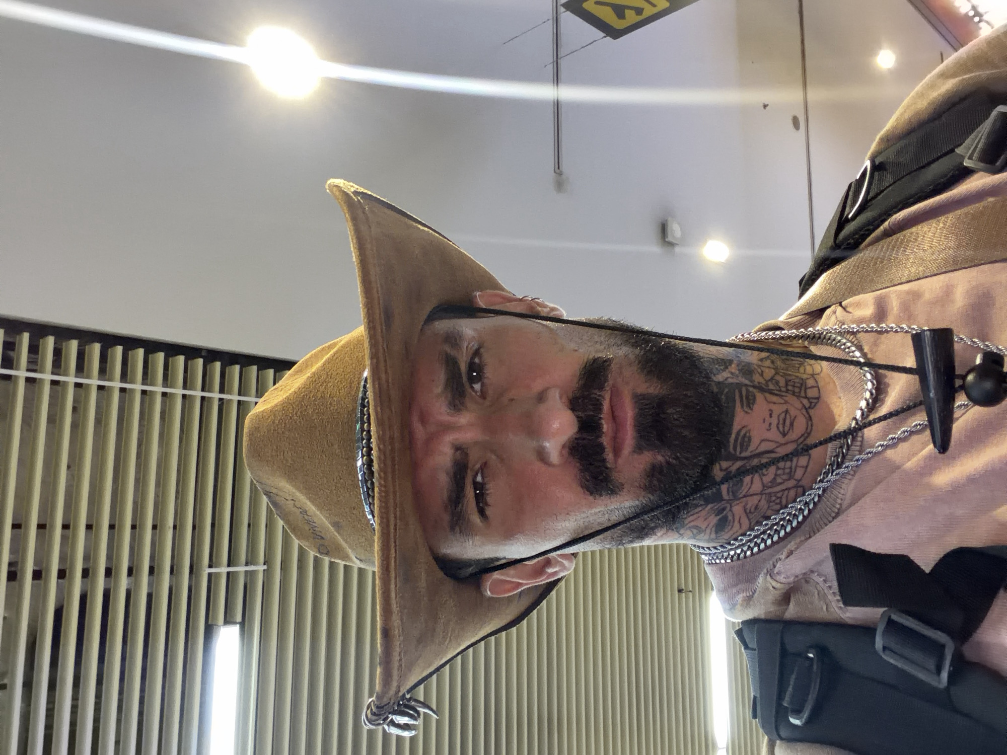 A medium shot photo shows a man wearing a brown cowboy hat and a pink shirt. He has tattoos on his neck and chest, and he is wearing several necklaces. He is also wearing a backpack. The background appears to be an indoor location with fluorescent lighting and a ceiling fan. The man's facial expression is serious, and he is looking directly at the camera. The overall impression is one of a casual, everyday scene.