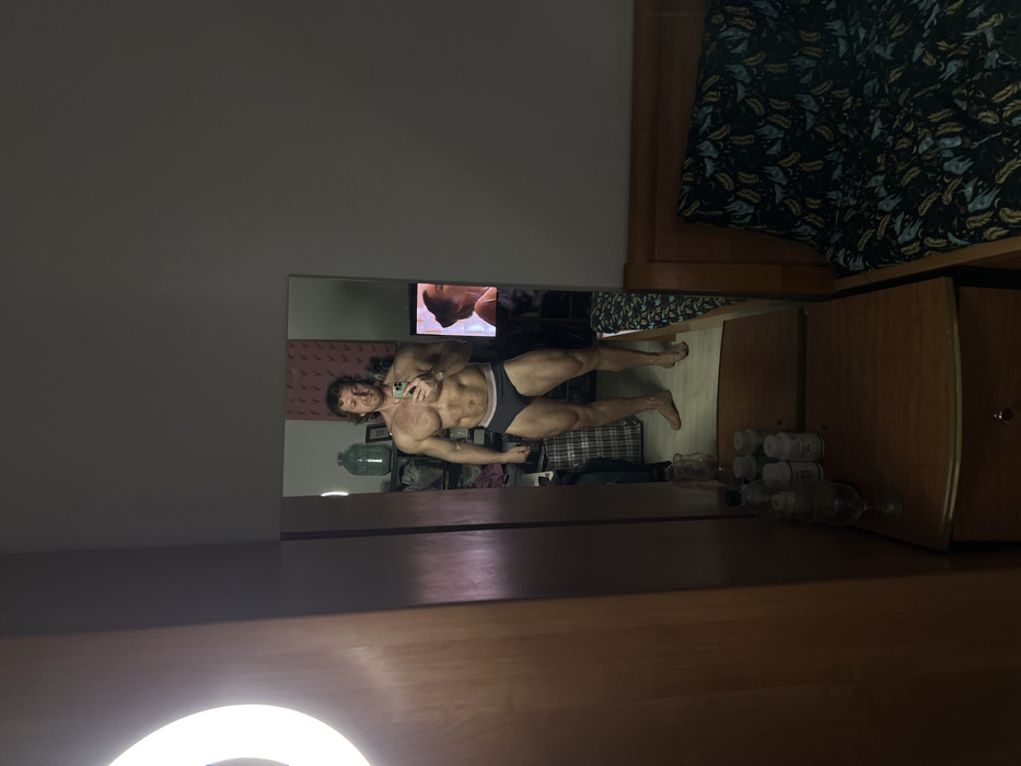 An indoor scene, stated to be located in Saint Petersburg, Russia, features a muscular adult male capturing a mirror selfie in what appears to be a bedroom or personal space. The man, with reddish-brown hair and a beard, stands barefoot, clad only in dark gray briefs, holding a light-colored smartphone to photograph his reflection.
Within the mirror's reflection, behind the man, is a light-colored wall adorned with a repeating pattern of reddish, antler-like shapes. A large, clear glass demijohn is visible to the left of the man's reflection, alongside an obscured collection of items resembling clothing or bags. To the right of the man's reflection, a television screen with a white bezel displays the profile of a woman with dark hair. The reflected floor is light-colored.
Outside the mirror's frame, to the left, a wooden wardrobe or cabinet is partially visible, with a prominent, brightly illuminated circular ring light attached. Directly below the mirror, a wooden nightstand with a single drawer holds several white plastic bottles, identified as supplements, and a clear glass containing liquid. To the right, a wooden bed frame is present, covered by a dark sheet or duvet featuring a pattern of lighter, possibly feather-like, shapes in green, blue, and yellow tones. The wall section to the right of the mirror is uniformly dark gray.