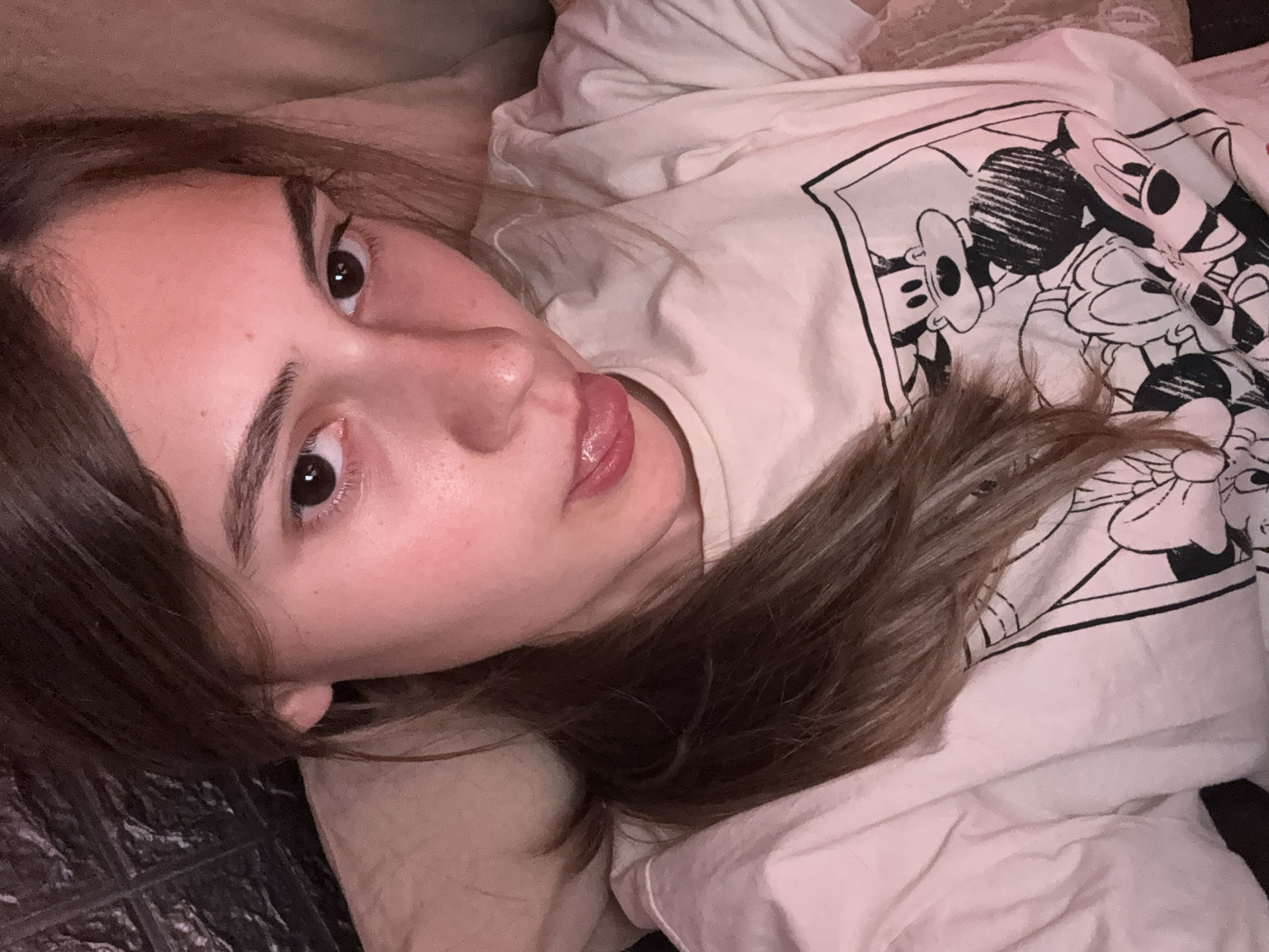 A young woman is depicted lying down in a close-up, slightly tilted shot, looking directly at the camera. Her head rests on a dark, textured surface, while her body is situated on a lighter, possibly off-white, soft surface, likely a bed or sofa. She has long, dark brown hair that cascades around her face and shoulders. Her eyes are dark and focused on the viewer. Her lips are slightly parted, with her tongue peeking out, conveying a relaxed or playful expression. Her complexion is fair and natural.
She is wearing a light-colored, possibly cream or white, t-shirt. The shirt features a distinct black and white graphic depicting cartoon characters, clearly identifiable as Mickey Mouse and Minnie Mouse, in a comic strip panel style. The fabric appears soft and comfortable.
The scene is indoors, with warm, soft lighting that suggests either evening or ambient artificial light. The atmosphere is casual and intimate. No specific location cues are visible in the image itself to indicate Panagyurishte, Bulgaria. There is no other legible text visible apart from the cartoon graphic. The overall emotion conveyed is one of ease and direct engagement with the camera.