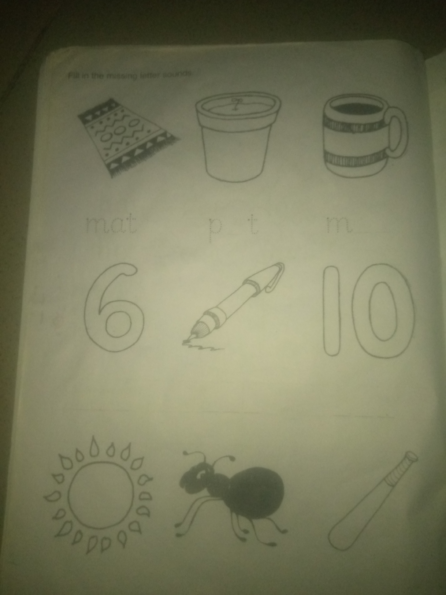 The image shows a page from a children's workbook with drawings and text. At the top, there's a title "Fill in the missing letter sounds". Below this are three sets of drawings. The first set shows a rug, a pot, and a mug. Underneath these are the words "mat", "p t", and "m". The second set shows the number "6", a pen, and the number "10". The third set shows a sun, an ant, and a baseball bat. The image is dimly lit and appears to be a scanned or photographed page, not a real-world scene. There is no information about the city of Jauro Yinu, Nigeria, or any forensic details related to a specific location or event.