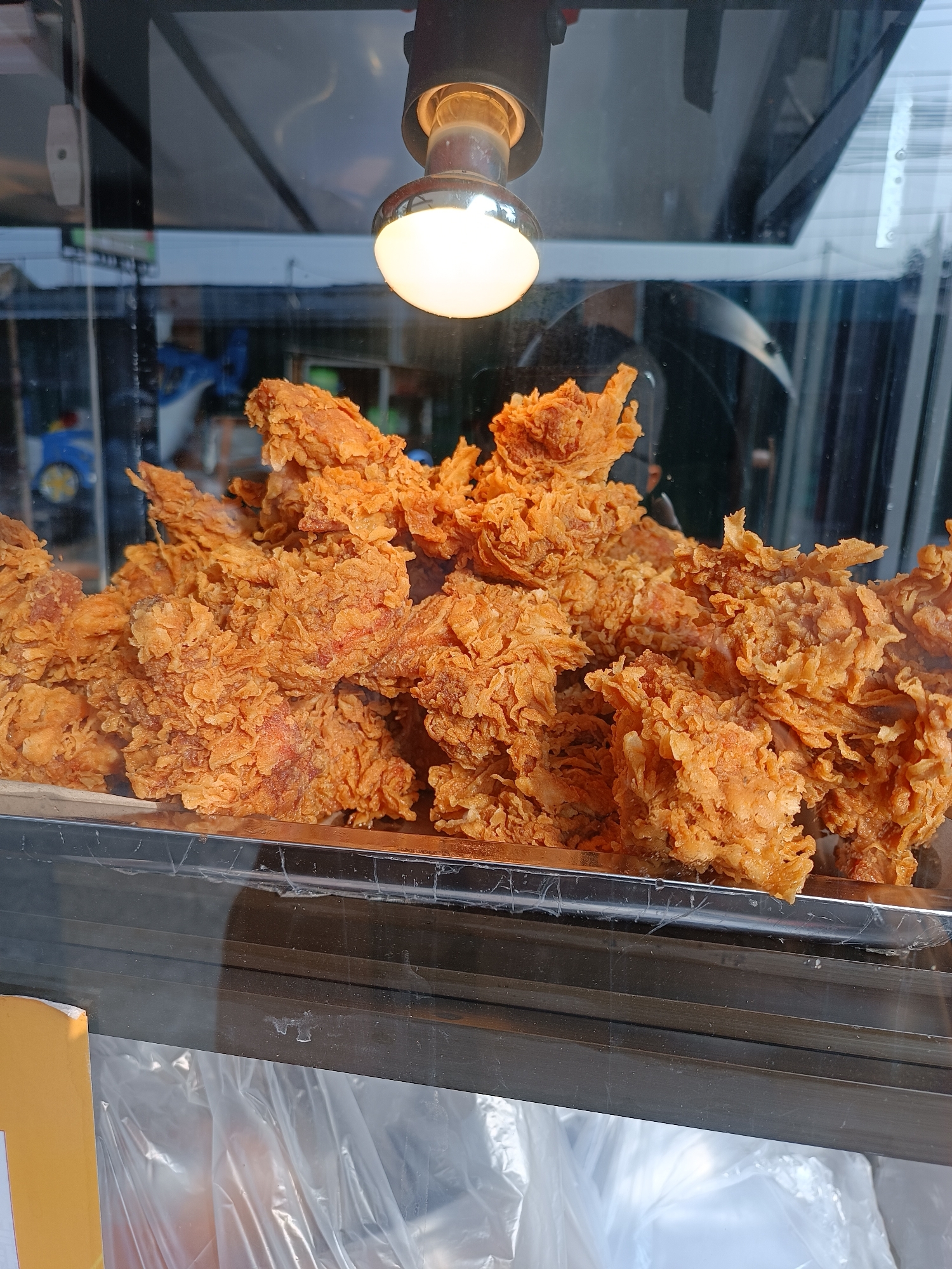 A close-up, eye-level shot shows a large quantity of deep-fried chicken pieces displayed in a metal tray. The chicken is golden brown and appears to have a crispy, textured coating. The tray is positioned behind a glass window, likely in a food stall or restaurant. Above the chicken, a bright light bulb illuminates the scene. Reflections are visible in the glass, showing a blurred background with hints of buildings and possibly vehicles. Below the tray, plastic bags are visible. The context provided indicates this scene is located in Imogiri, Indonesia, suggesting it is likely a street food vendor or a local eatery.