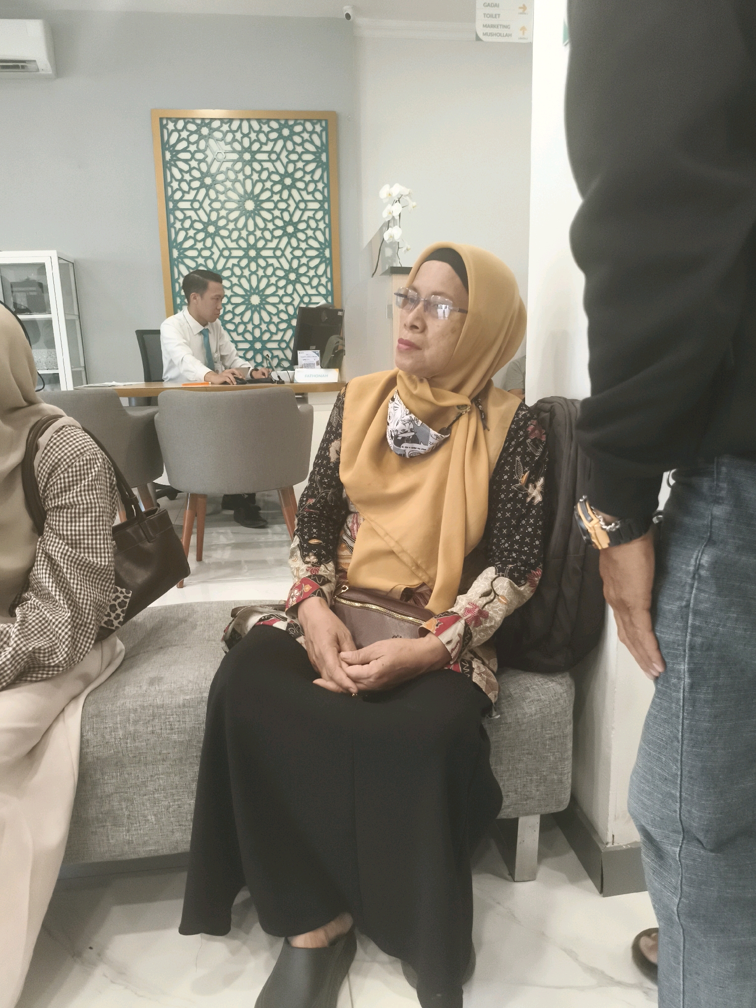 In a well-lit indoor space that appears to be a financial institution or office lobby in Turikale, Indonesia, three individuals are visible. A man in a white collared shirt and a blue tie is seated at a wooden desk in the background, facing a computer monitor. To his left, a decorative patterned screen adorns the wall.
In the foreground, a woman wearing a mustard-yellow hijab and glasses is seated on a grey upholstered bench. She is dressed in a black long-sleeved top with a patterned fabric on the sleeves and a black skirt. A brown clutch bag rests on her lap, and her hands are clasped together. To her left, another person is partially visible, wearing a beige hijab and a patterned long-sleeved top. A dark brown handbag with a textured detail hangs from this person's shoulder.
To the right of the seated woman, a person wearing a black long-sleeved garment and blue jeans stands facing away from the camera, obscuring a portion of the background. The floor is tiled with a white marble pattern. The environment suggests a formal setting, possibly a waiting area or customer service point.