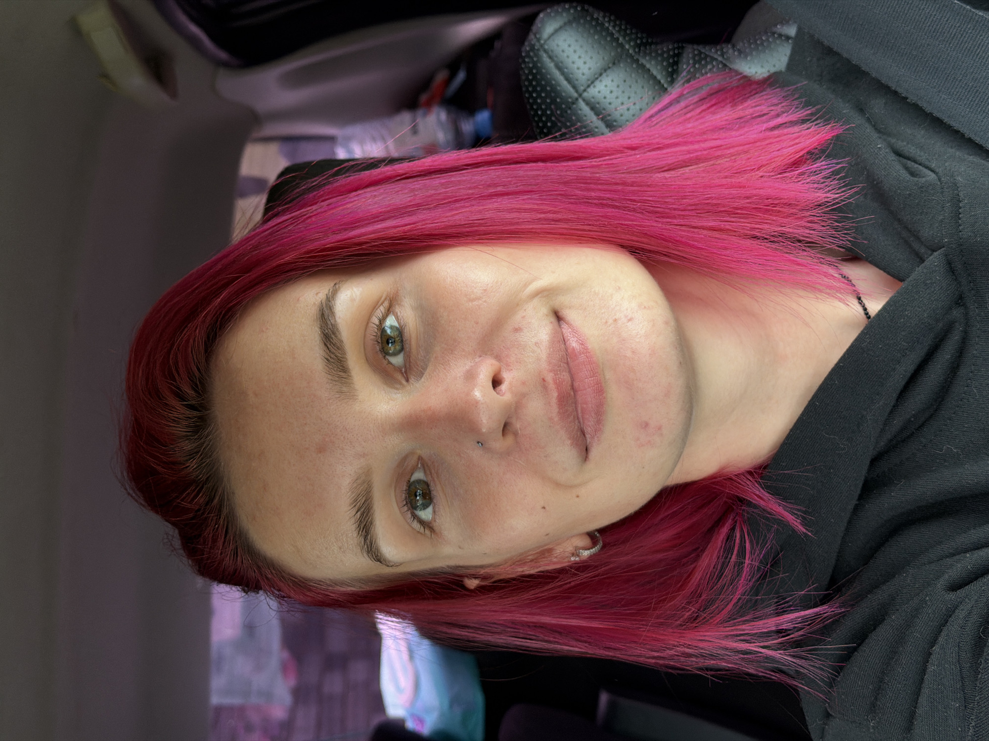 A young woman with bright pink hair sits in the passenger seat of a car. Her head is slightly tilted and she is looking towards the camera with a subtle smile. She has a nose piercing and a hoop earring in her left ear. The background suggests an urban environment, possibly within the city limits of Chaikovsky, Russia, with blurred buildings and a hint of construction or industrial activity visible through the car window. The interior of the car is visible, including a black quilted seat. The lighting is natural, coming from the front, casting soft shadows on her face.