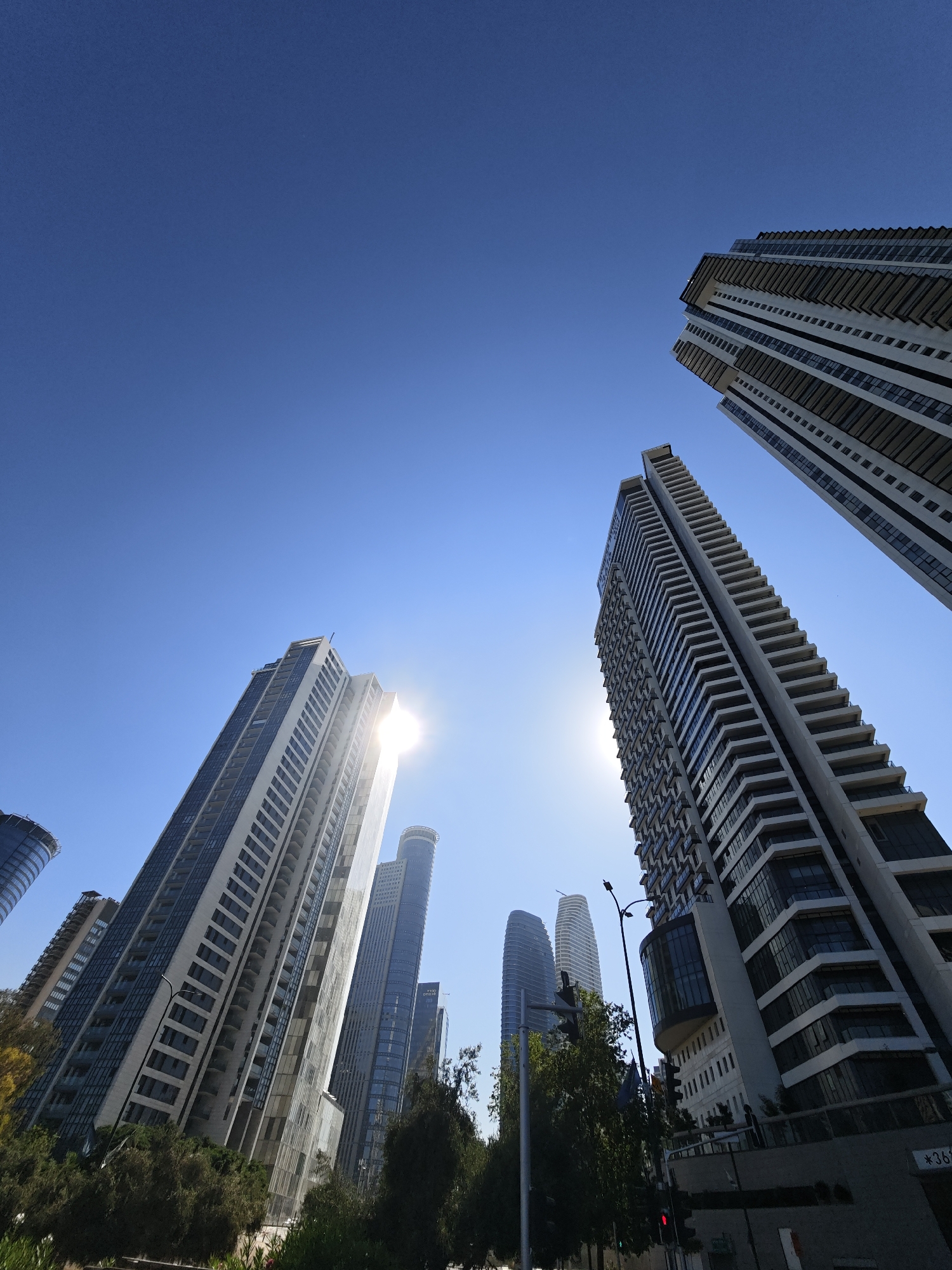 This is a low-angle, wide shot of a modern urban cityscape dominated by a cluster of tall, contemporary skyscrapers. The buildings, varying in height and architectural style, are made of glass and concrete, featuring numerous balconies and sleek facades. The sun is shining brightly, creating a lens flare effect in the upper center of the image, and casting a slight glare on some of the buildings. The sky is a clear, vibrant blue. In the foreground, lush green trees and foliage line a street, suggesting a well-maintained urban environment. A traffic light and a street lamp are visible, indicating an active street scene. The architecture suggests the location is likely a business or residential district in a developed city. The presence of the "ISS" logo on one of the buildings, and the overall architectural style, points to the city of Givatayim, Israel.