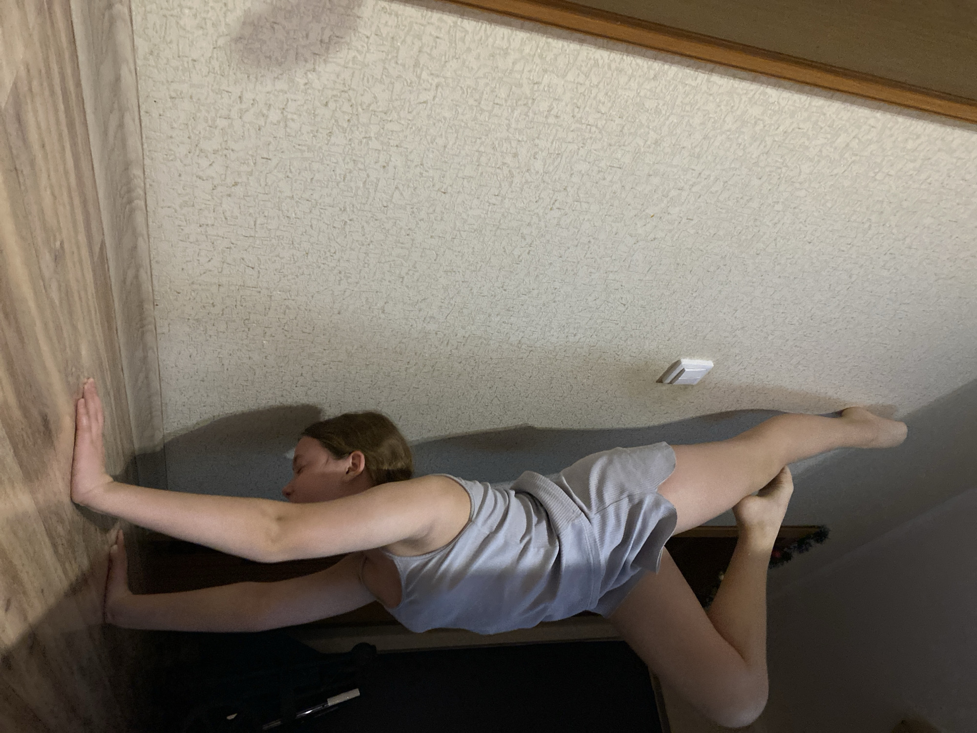 A woman in a gray top and shorts is performing a yoga pose, possibly a standing bow pose, with her hands pressed against a light brown wooden wall. She is in an interior space with a textured off-white ceiling featuring a wooden trim.