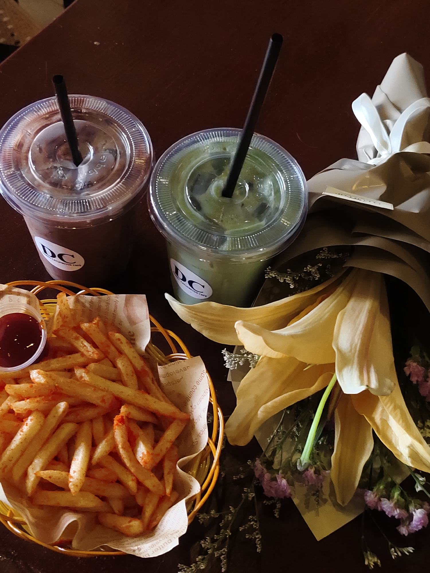 The image depicts a table setting at a cafe, likely in Davao City, Philippines, featuring two iced beverages in clear plastic cups with black straws, a small basket of french fries with a side of ketchup, and a bouquet of yellow lilies with smaller purple flowers. The drinks appear to be a chocolate beverage and a green tea matcha latte, both with the "DC" logo visible on the cups. The french fries are golden brown and seasoned, served in a yellow woven basket lined with white paper. The bouquet is elegantly wrapped in brown paper and white ribbon. The dark brown table surface suggests an indoor setting. There are no people present in the frame.