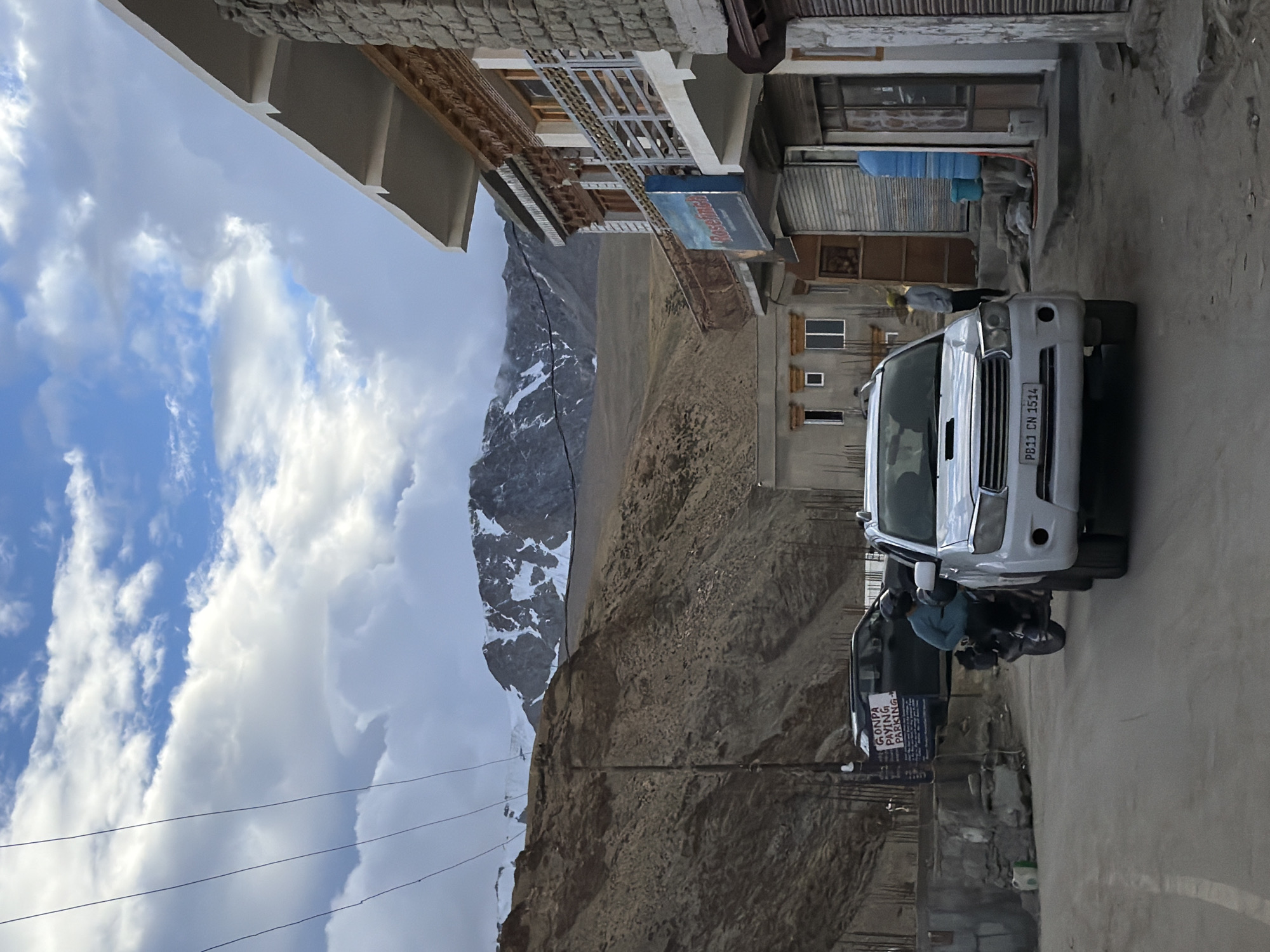 A white SUV, license plate PB11 CN 1514, is parked on a dirt road in Karzok, India. To its right, a building with intricate wooden carvings and a balcony is visible. A man in a yellow turban stands near the building entrance. To the left of the SUV, a motorcycle with a rider wearing a blue jacket and helmet is moving away. Behind the motorcycle, a dark SUV is parked near a sign that reads "GONPA PAYING PARKING". In the background, barren mountains with snow-capped peaks are visible under a cloudy sky. The scene appears to be a street in a high-altitude village.