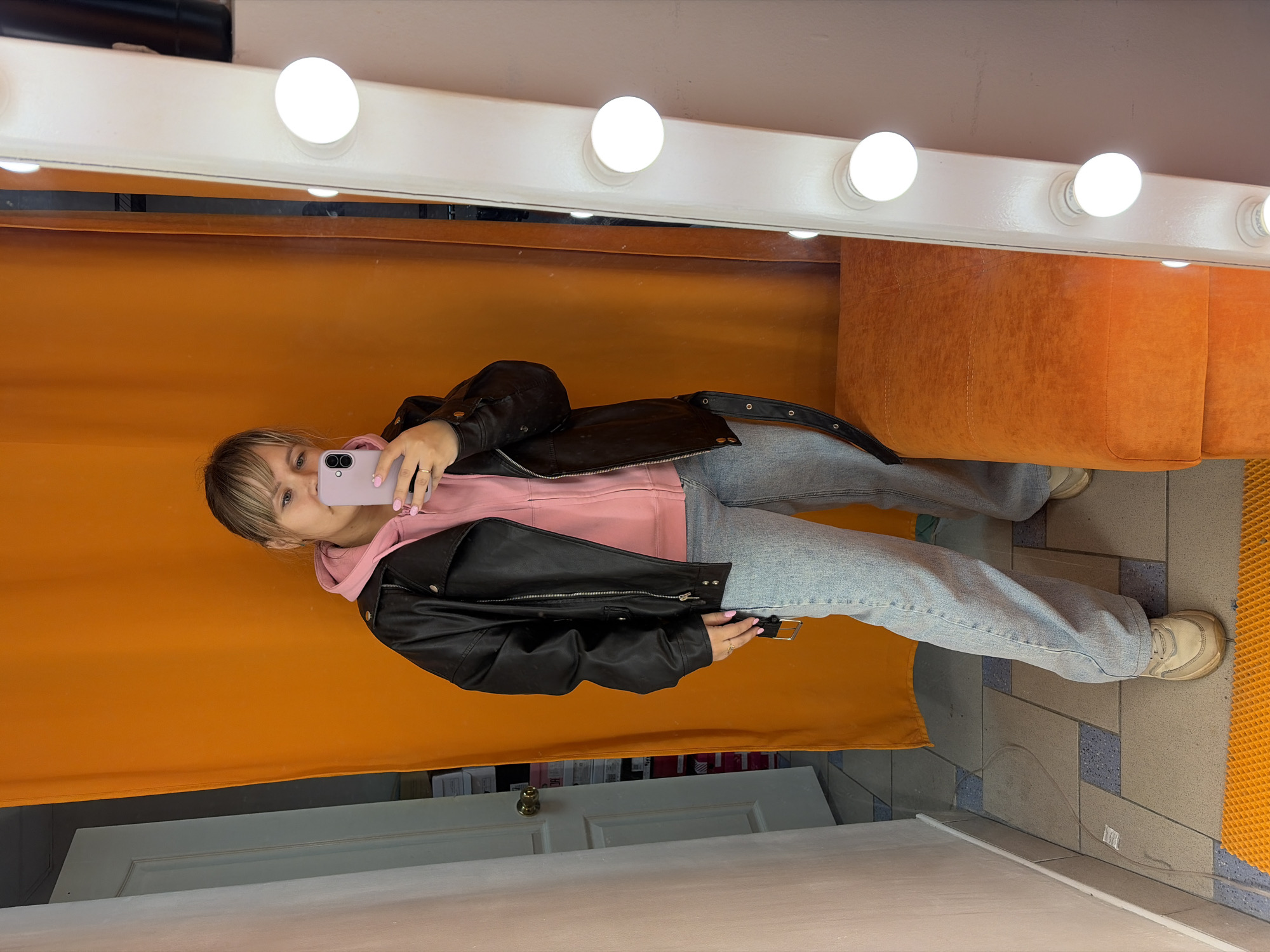 The image captures a young woman taking a mirror selfie in what appears to be an indoor dressing room or vanity area. She is holding a light purple smartphone with a prominent camera array, angled to capture her reflection.
The woman is wearing a casual outfit consisting of a pink hooded sweatshirt, a black faux leather jacket adorned with metal zippers and snaps, light-wash blue jeans, and light-colored sneakers. A ring is visible on her right hand.
The setting is characterized by a large mirror, which dominates the scene. Along the top edge of the mirror, a strip of several evenly spaced, illuminated round bulbs provides bright, direct lighting. In the reflection, behind the woman, there is a vibrant orange curtain or a fabric-covered wall. An orange upholstered bench or seating unit is also visible in the reflection to her right. The floor is tiled with large, rectangular grey and blue patterned tiles, and a section of a textured orange mat is visible on the far right. Below the woman's reflection, a white door with a brass doorknob is reflected in the mirror.
The activity is clearly a self-portrait taken using a phone reflection. The mood is neutral, focused on the act of photography. There is no visible text that can be clearly deciphered.