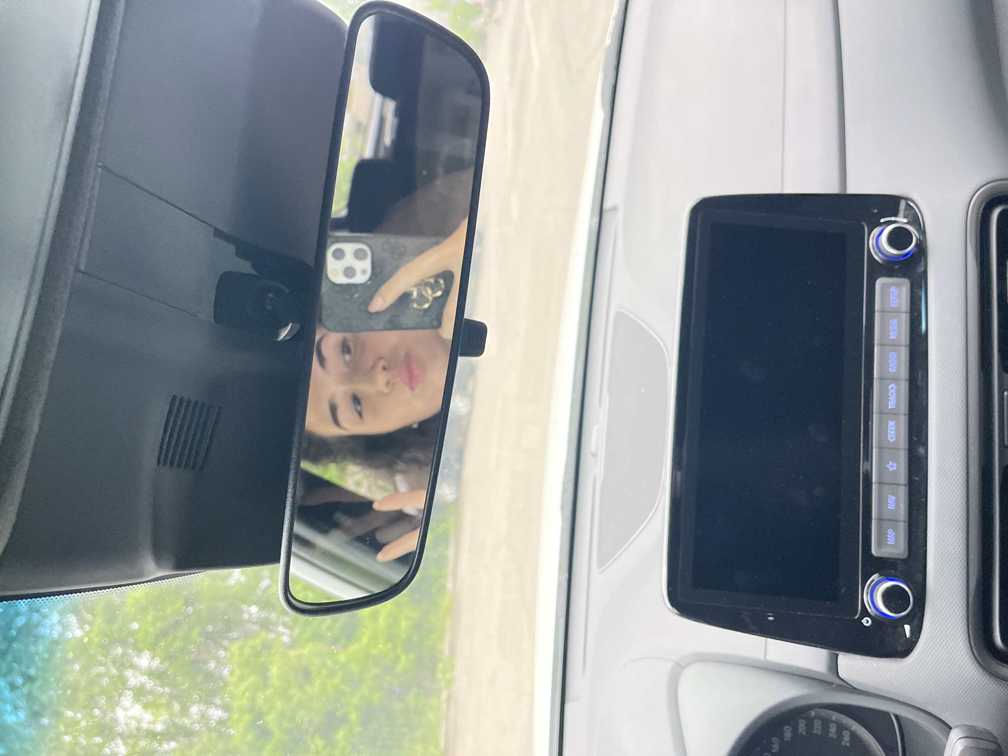 The image captures an interior view of a car, likely situated in Sofia, Bulgaria, during daytime. The primary subject is a person taking a selfie, reflected in the rearview mirror. The reflection shows a female with dark, curly hair and light-colored lips, holding a smartphone with a triple-lens camera system and a dark, patterned case. Her pose suggests a casual, perhaps playful, expression.
To the right of the rearview mirror, a segment of the car's dashboard and infotainment system is visible. The infotainment unit features a dark display screen and several control buttons, some illuminated with blue light. Text visible on these buttons includes "MAP," "NAV," "SEEK," "TRACK," "RADIO," "MEDIA," and "SETUP." A rotary knob is also present, likely for volume or power. Below this, a portion of the instrument cluster or speedometer is seen, displaying numerical values such as "160," "180," "200," "220," "240," and "260."
Through the car's windshield and the upper part of the driver's side window, bright green foliage and diffuse sunlight are apparent, indicating bright, outdoor conditions.