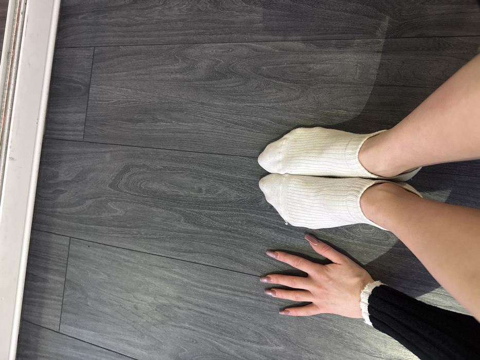 A person wearing white socks is seen from an overhead perspective, on a grey wood-patterned floor. Their legs are angled towards the right of the image. A hand with light skin and elongated nails rests on the floor. The person is wearing a black garment with a white frilled edge on their wrist. A white frame is partially visible on the left edge.