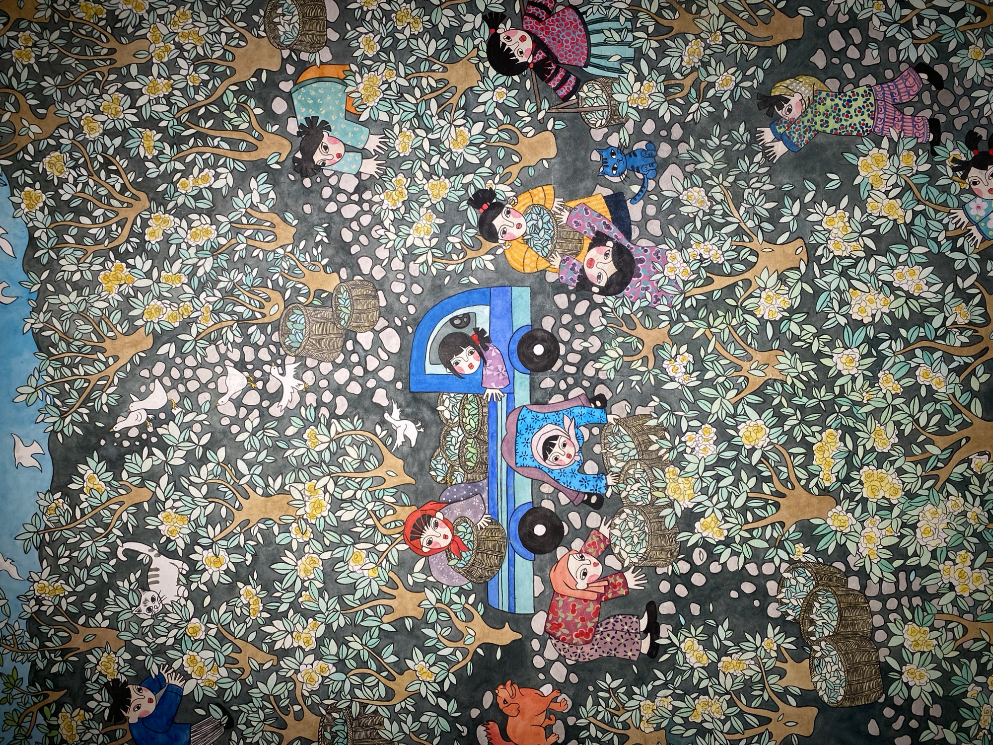 This is a colorful, densely packed mural depicting a harvest scene in a garden, set against a dark, textured background. The mural is populated by numerous stylized human figures, predominantly children, and several animals. The human figures are rendered in a flat, graphic style with bold outlines and vibrant clothing patterns. They are shown engaged in activities related to harvesting, such as picking fruit or vegetables, carrying baskets, and loading them onto a blue truck. The children have distinct facial features with large eyes and expressive mouths.
Animals present include a ginger-colored dog on the left, a white cat near the top left, several white birds scattered throughout, and a small blue cat sitting near the bottom center. The environment is characterized by numerous trees with brown trunks and branches, adorned with clusters of small yellow and white flowers and green leaves. The ground is covered with light-colored, rounded stones.
The mural appears to be located on a wall in Yuzhong District, China, given the context provided. The overall impression is one of a busy, communal harvest, with a playful and slightly whimsical atmosphere. The composition is layered and busy, with figures and natural elements interwoven throughout the frame. The lighting appears to be even, with no strong shadows, suggesting it is an indoor mural or lit artificially.