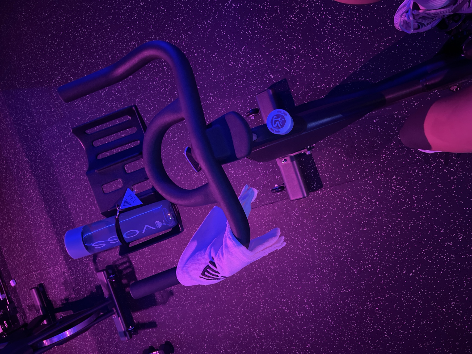 The scene presents an indoor cycling bike situated on a purple-toned, textured floor. The bike features a water bottle in a holder, a towel draped over the handlebars, and a visible tension control knob. A person's leg and shoe are partially visible to the upper right. The image is taken in a setting with strong purple lighting. No city or location is apparent.