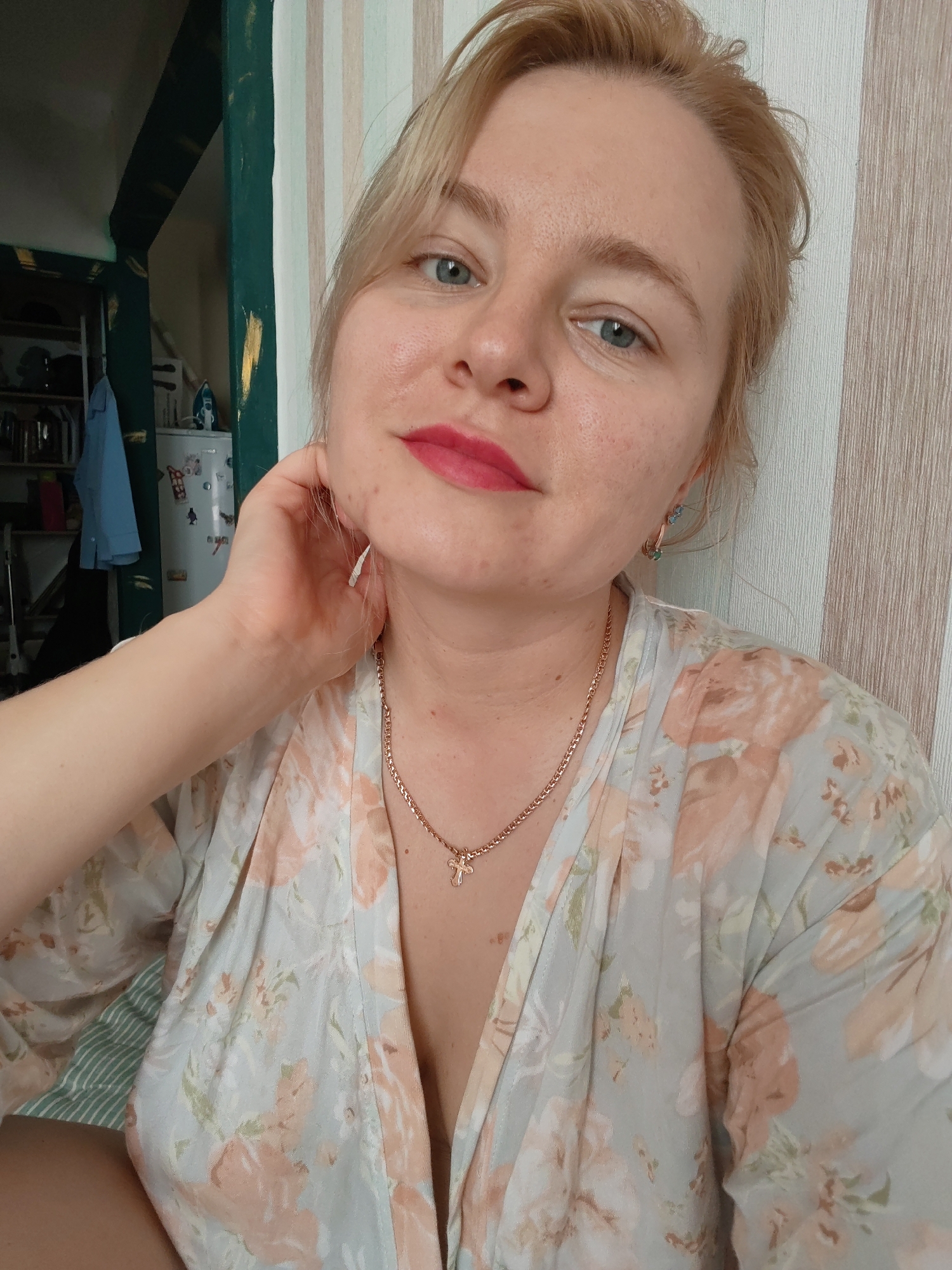 A medium shot shows a woman with blonde hair and blue eyes, wearing a floral robe. She is touching her neck with her right hand and looking at the camera. She has bright pink lipstick on. Behind her is a wall with vertical stripes and a shelf unit with various items. The environment appears to be indoors, likely a residential setting. The image appears to have been taken in Izhevsk, Russia.