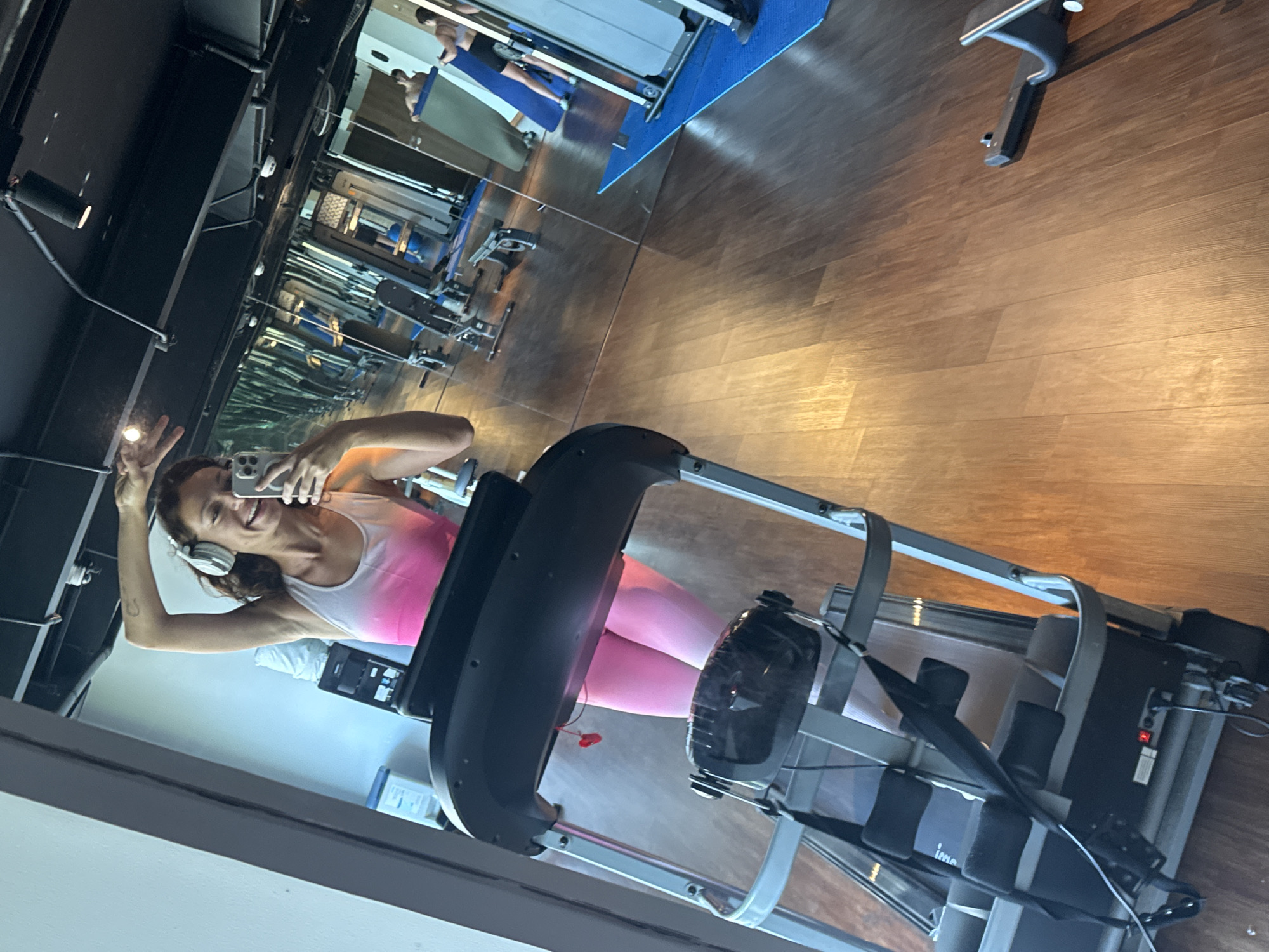 The image shows a woman in a pink and white tank top and headphones taking a selfie in front of a mirror in a gym. She is on a rowing machine. Reflected in the mirror are other gym machines and people exercising. The gym appears to be well-equipped and has a wooden floor. The location appears to be in a mirrored gym. The gym may be in Đà Nẵng, Vietnam.