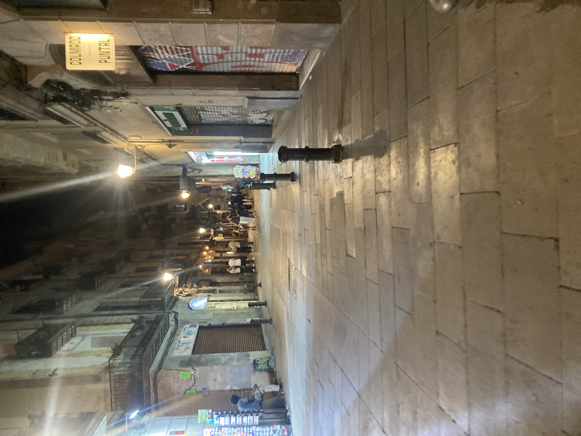 This image depicts a narrow street in Barcelona, Spain, at night. The street is paved with light-colored rectangular stones. Lining the left side of the street are old buildings with balconies and various shops with awnings. A sign reading "Colmado Puntal" is visible. Several dark cylindrical objects are placed along the edge of the street on the right, casting long shadows from the streetlights. In the distance, there are blurred figures of people walking and dining at outdoor tables, illuminated by streetlights. The overall atmosphere is dimly lit and suggests a lively urban scene.
