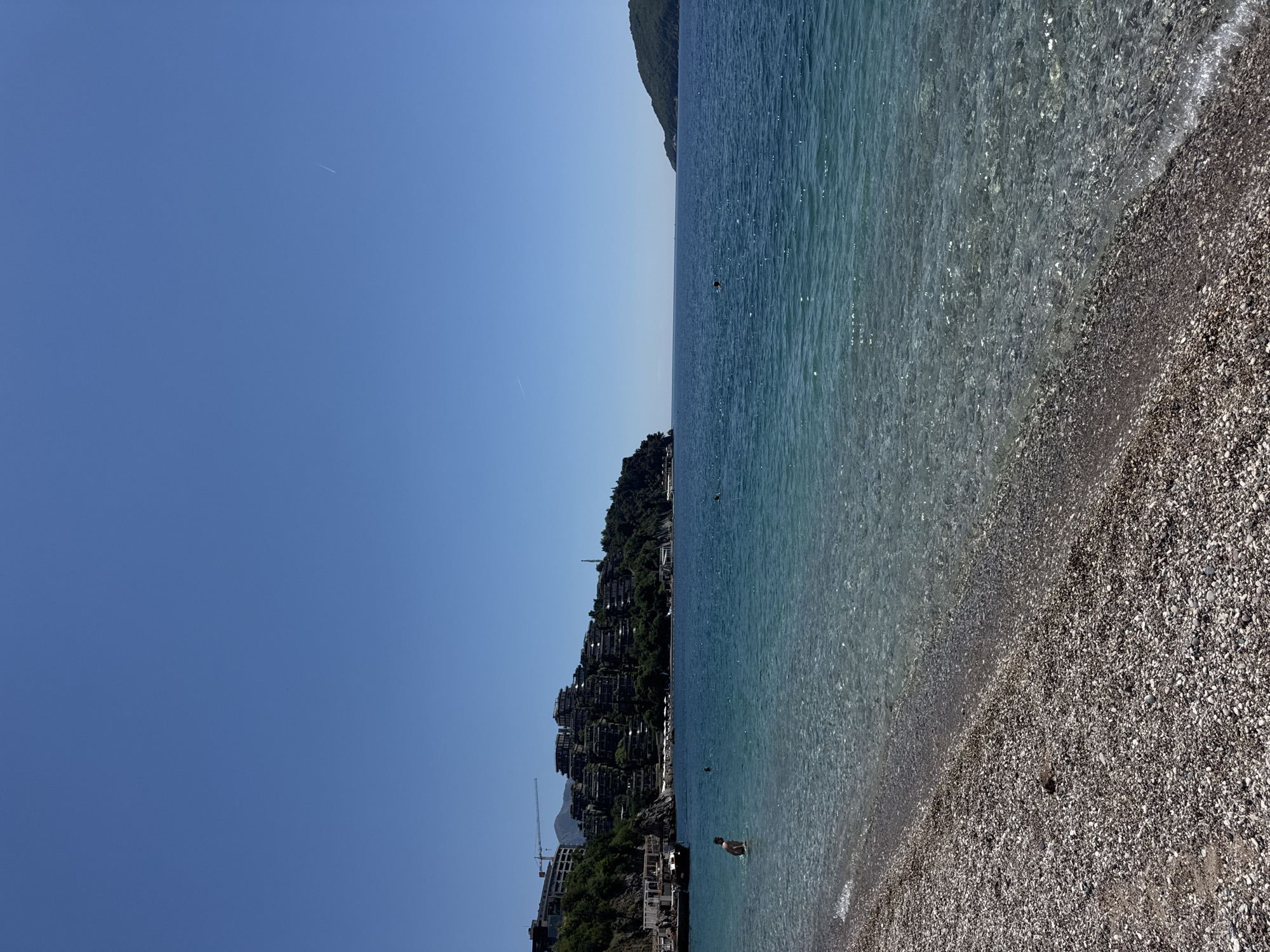 The image depicts a beach scene in Budva, Montenegro. The view is oriented vertically. To the right is the Adriatic Sea, which is clear and turquoise, transitioning to a darker blue further out. The shoreline consists of small grey pebbles. To the left, a steep, green hillside supports a complex of modern buildings, possibly apartments or a resort. A construction crane is visible among the buildings. The sky is clear and bright blue. There is a contrail high in the sky. A few people are visible in the water.